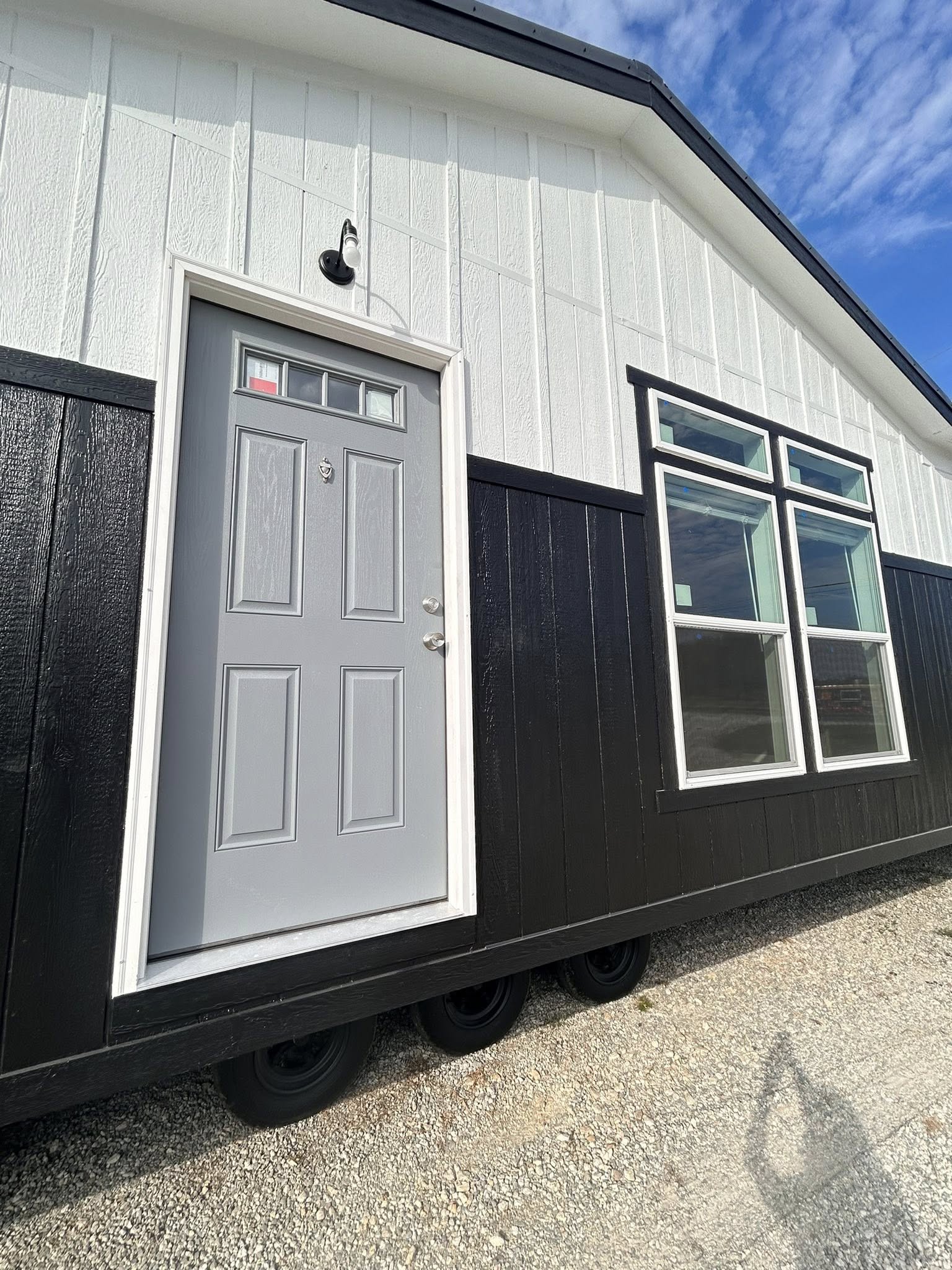 Modern tiny house exterior with a black and white facade, featuring a gray door and large windows. Wheels underneath, set against a bright blue sky.