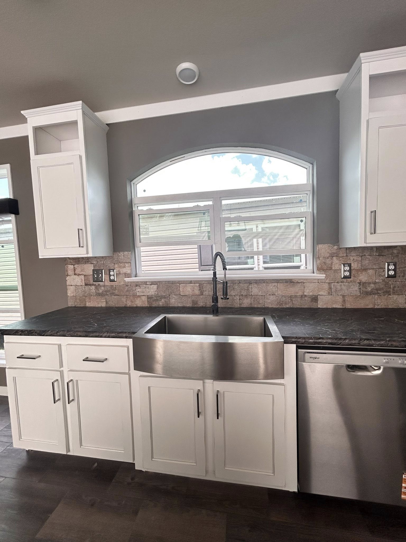 Modern kitchen with white cabinets, stainless steel sink, and dark countertops. A large arched window above provides natural light. Cozy and clean ambiance.