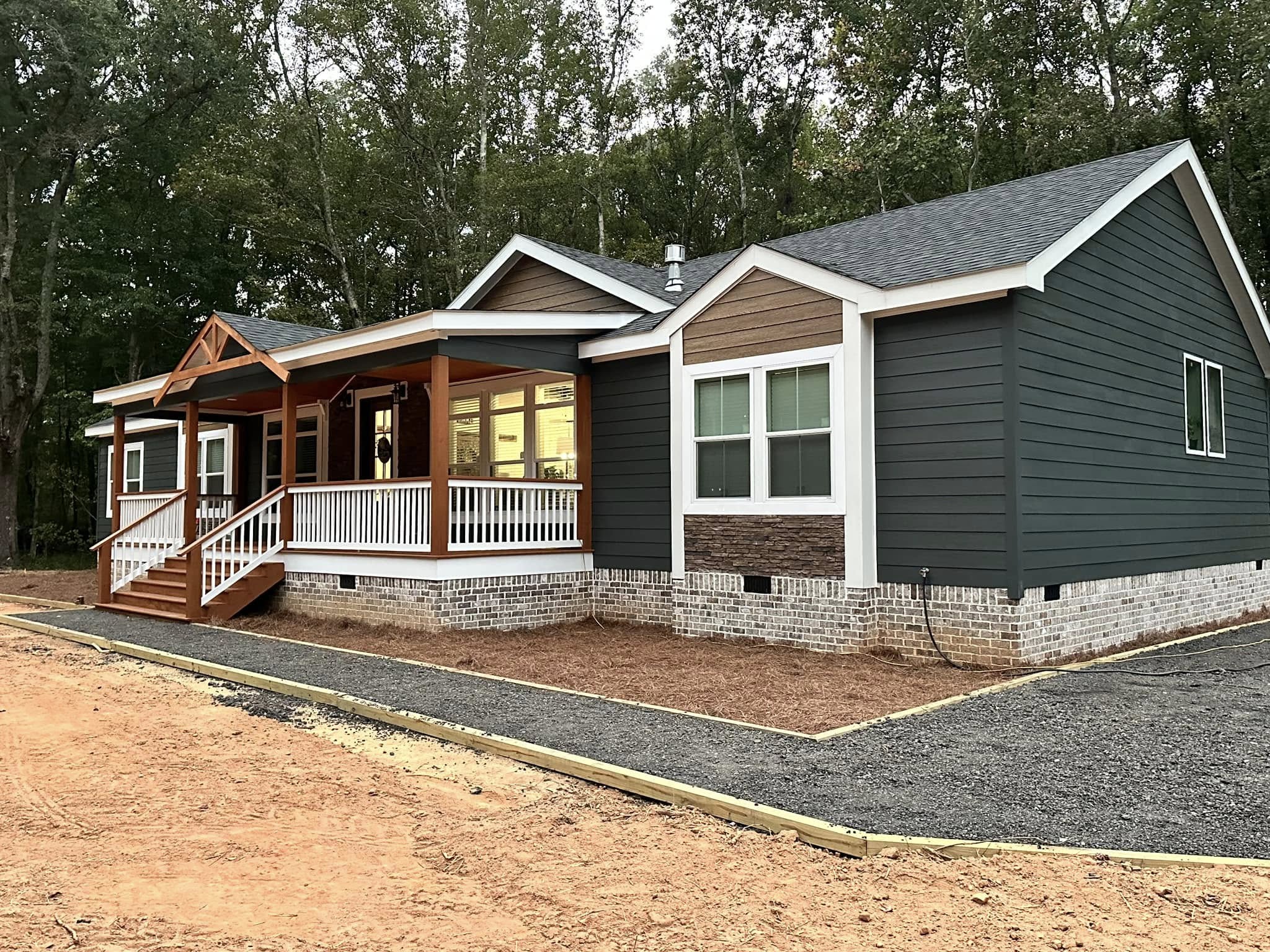 A modern, dark gray ranch-style house with a white-trimmed porch and brick foundation, set against a backdrop of tall trees, conveying a cozy, rustic feel.