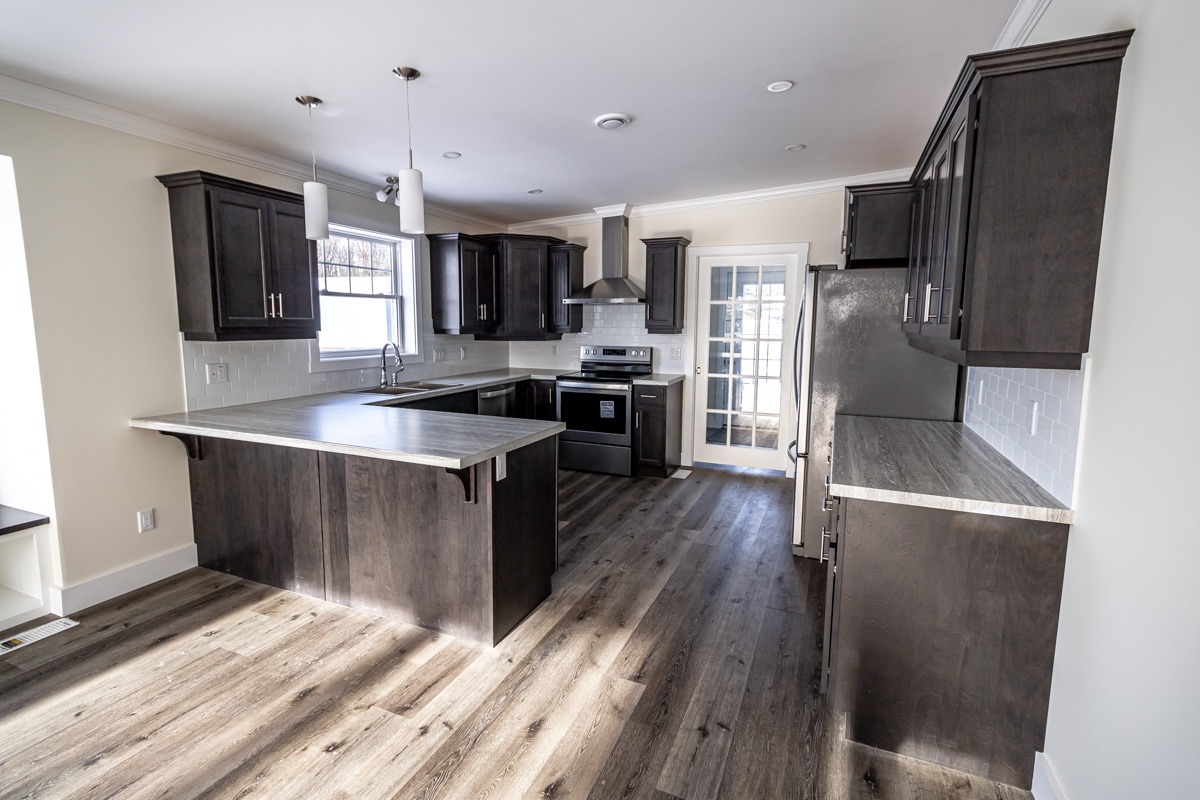 Modern kitchen with dark wood cabinets, light countertops, stainless steel appliances, and wood flooring. Sunlight enters through a window and glass door.