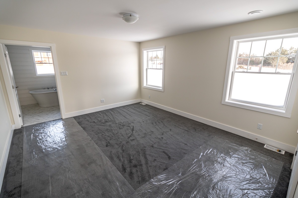 Empty room with freshly installed gray carpet partially covered by plastic, beige walls, and two windows providing natural light. A doorway leads to a white-tiled bathroom.
