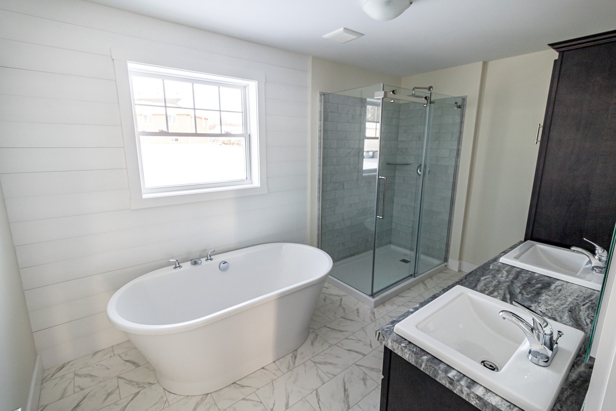 Modern bathroom with a white freestanding tub under a window, glass-enclosed shower, and dual sinks with granite countertop on tiled floor. Bright and clean.