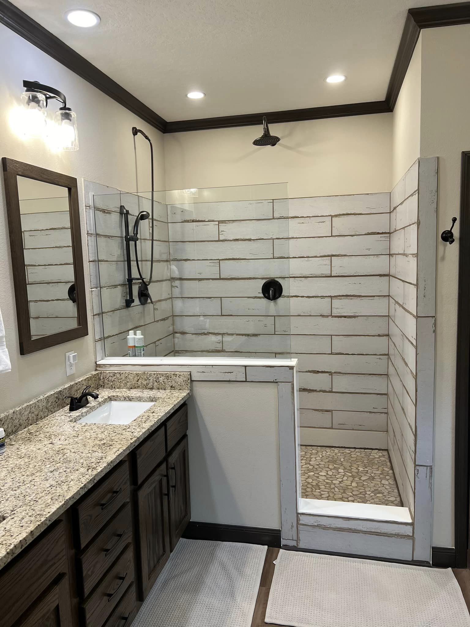 Modern bathroom with rustic, whitewashed wood-plank tiles in the shower. Granite countertop with wooden cabinets and lit mirror. Cozy, serene feel.