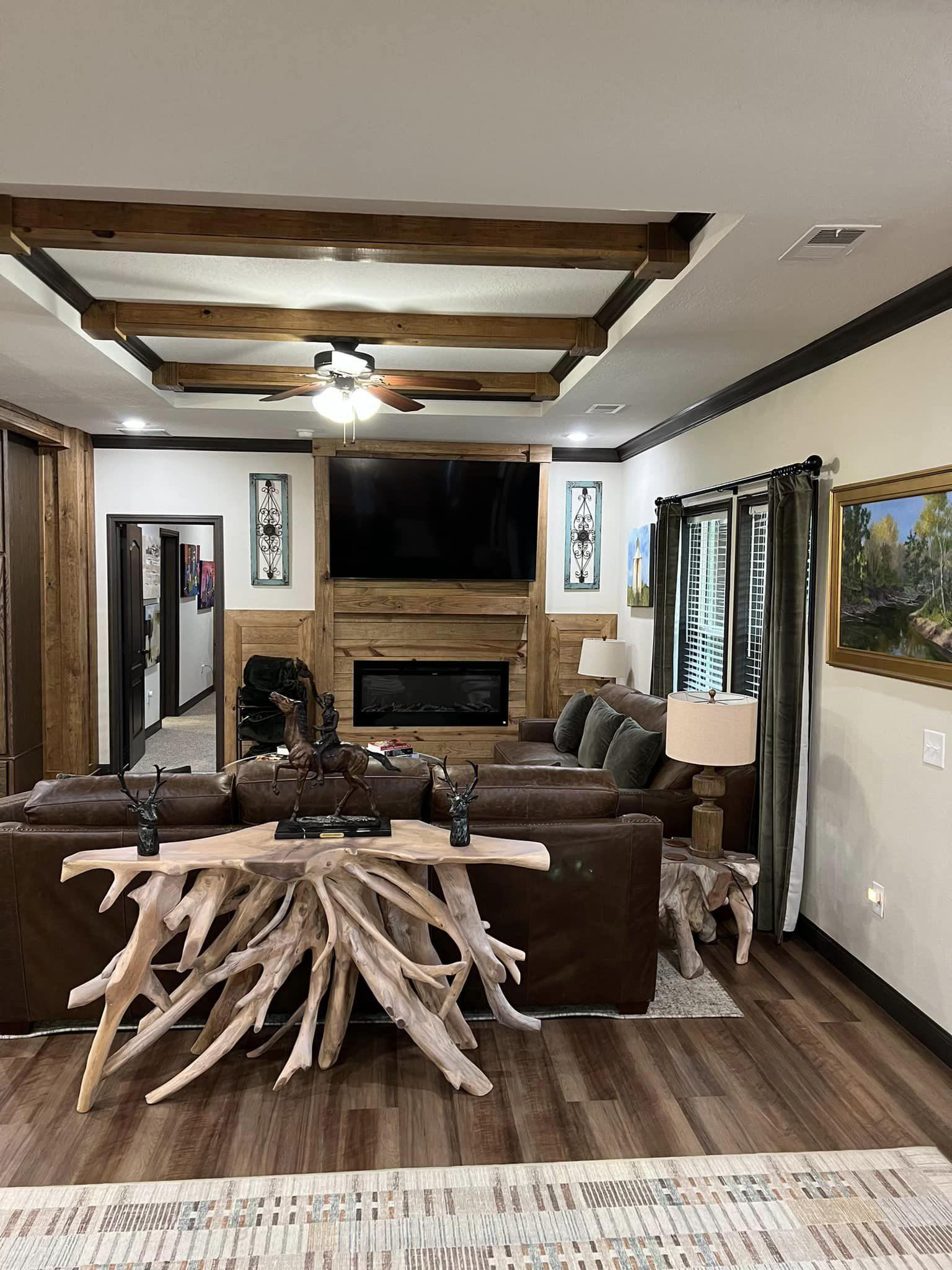 Cozy living room with wood accents, featuring a large TV above a fireplace, leather sofas, a driftwood coffee table, earth-tone decor, and landscape paintings.
