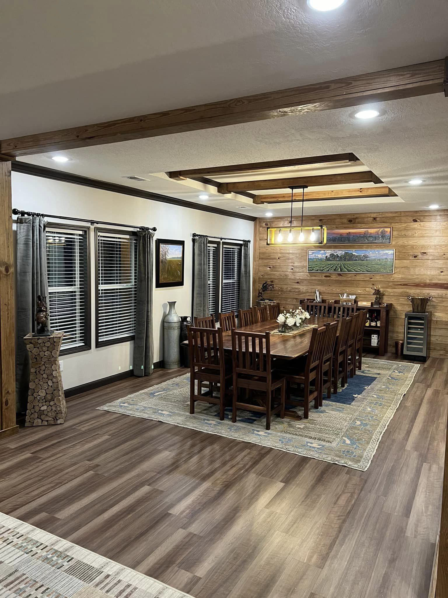 Warm, rustic dining room with a long wooden table and chairs set for twelve. Wooden beams, cozy lighting, and wall art enhance the inviting atmosphere.