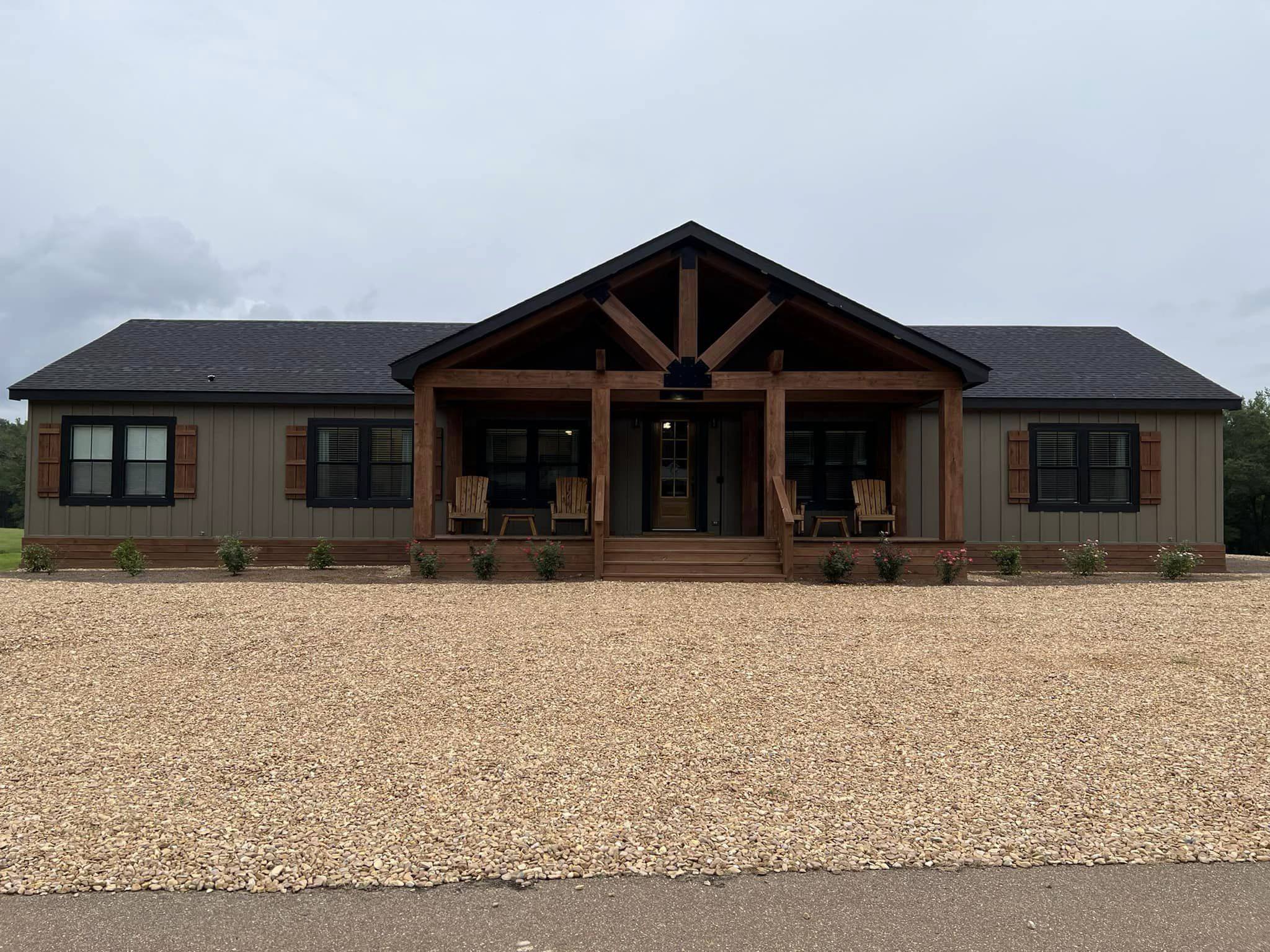 Single-story house with rustic wooden porch and rocking chairs, surrounded by a gravel yard. Cloudy sky. Calm, inviting rural setting.