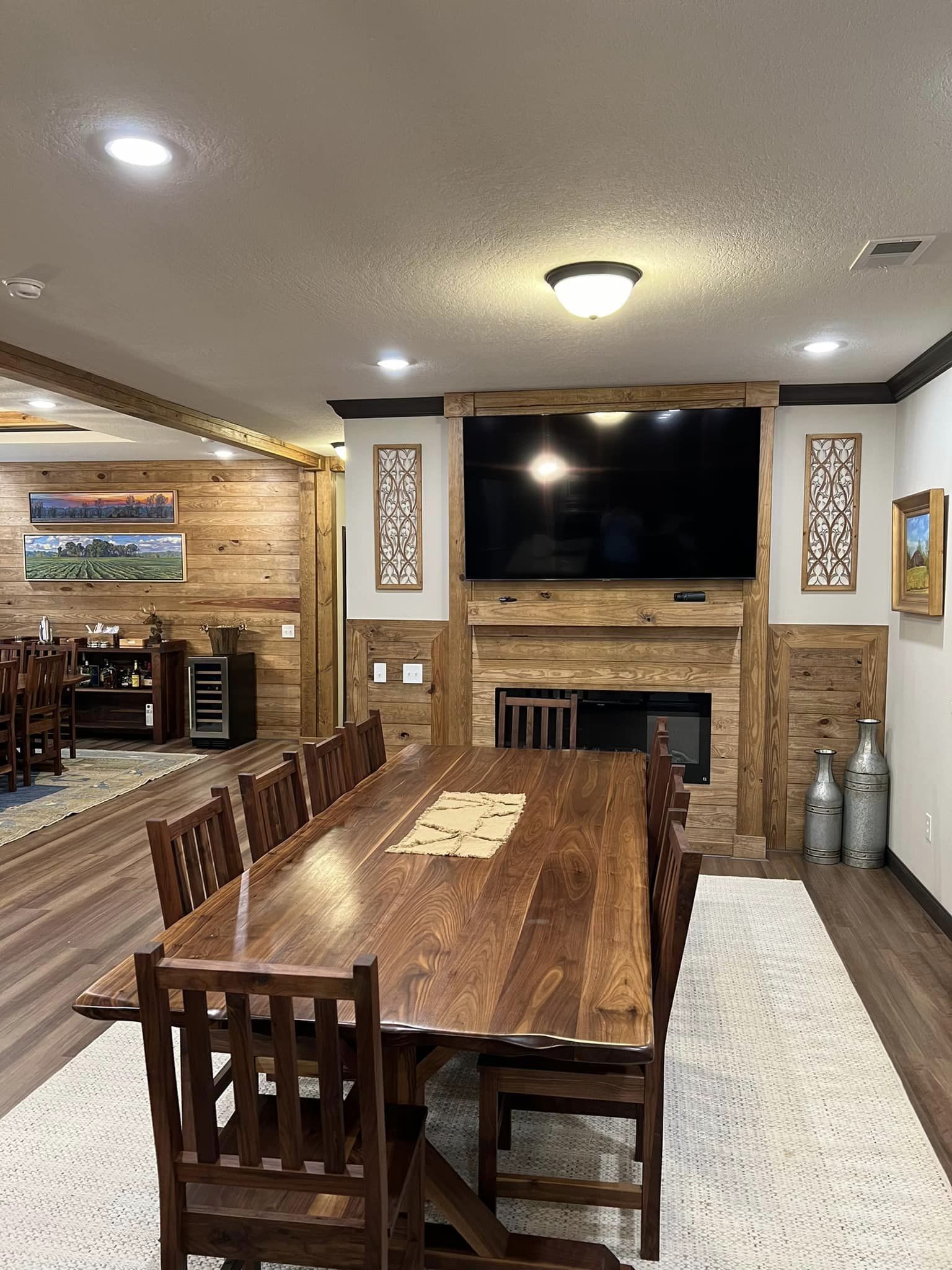 Rustic dining room with a long wooden table, eight chairs, and a large TV above a wood-paneled fireplace. Warm lighting and decorative wall art.