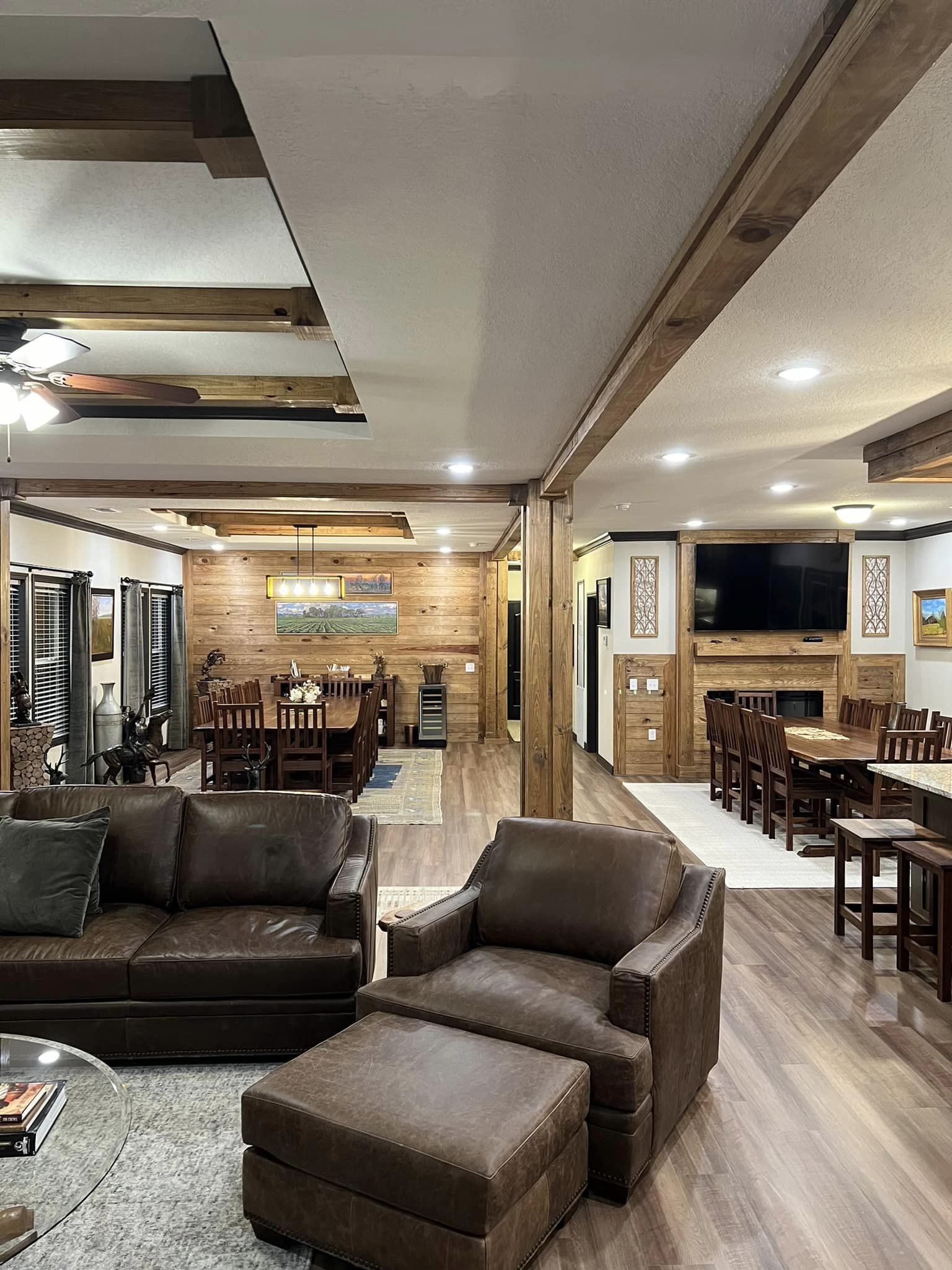 Spacious living area with brown leather sofa and armchair, wooden beams, and warm lighting. Features dining table, wooden walls, and flat-screen TV.