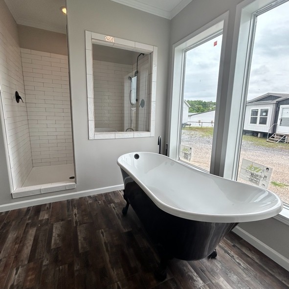 A modern bathroom with a black freestanding tub on dark wood floors, next to a white-tiled shower. Large windows offer an outdoor view.
