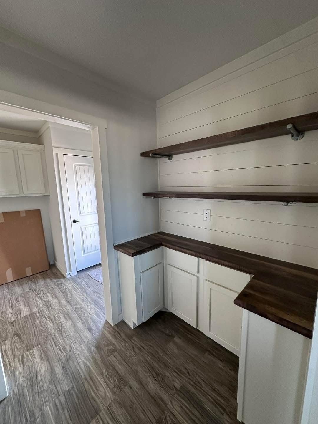A small, modern pantry with white cabinets and dark wooden countertops and shelves. The light wood flooring complements the bright, minimalist decor.