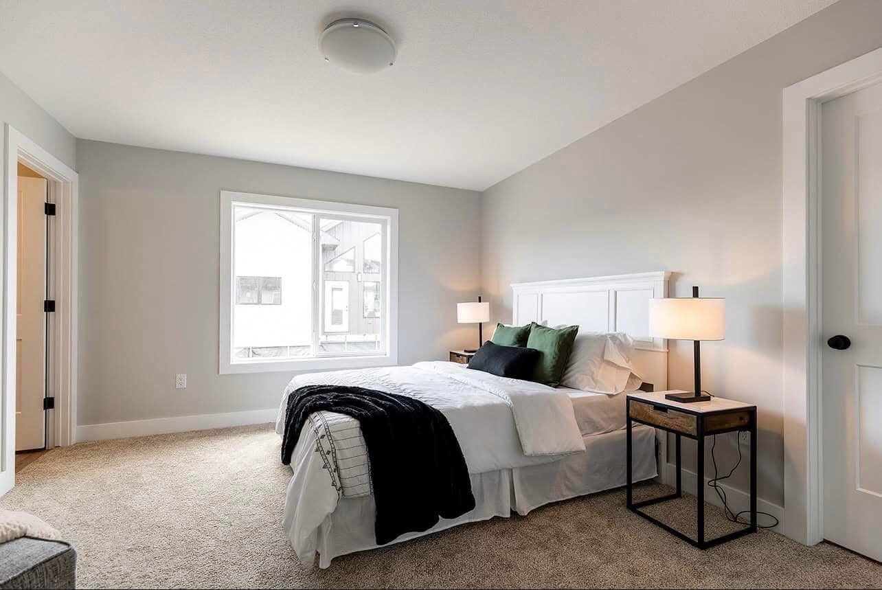 A modern bedroom with a neatly made bed featuring white linens and green and black pillows. A black nightstand with a lamp is beside it. Large window with natural light. Neutral tones and cozy atmosphere.