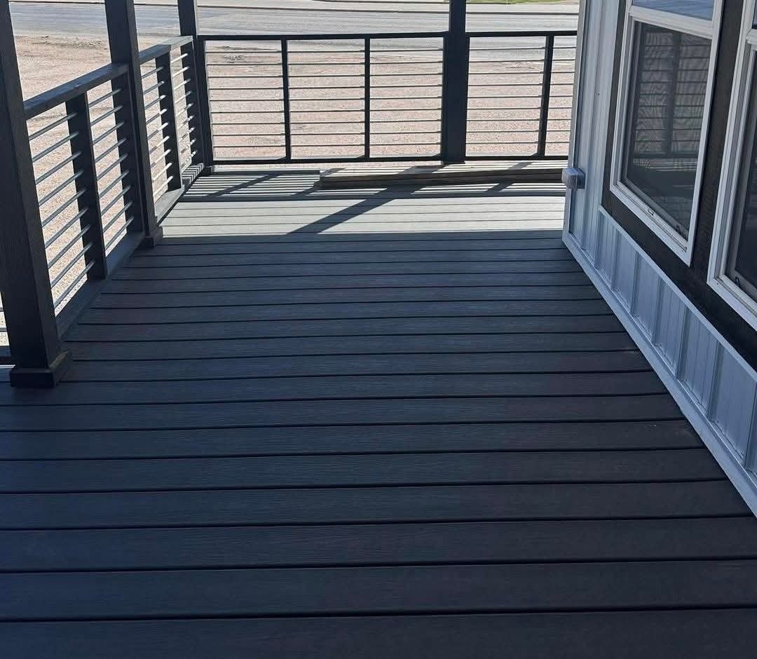 A wooden deck extends from a house with large windows on the right. Sunlit railings cast shadows across the gray planks, creating a tranquil outdoor space.