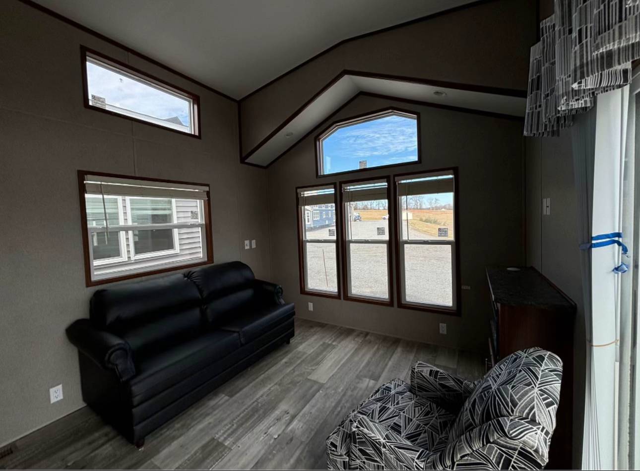 Modern living room with dark sofa, patterned armchair, large windows, and hardwood floor. Natural light fills the space, creating a cozy ambiance.