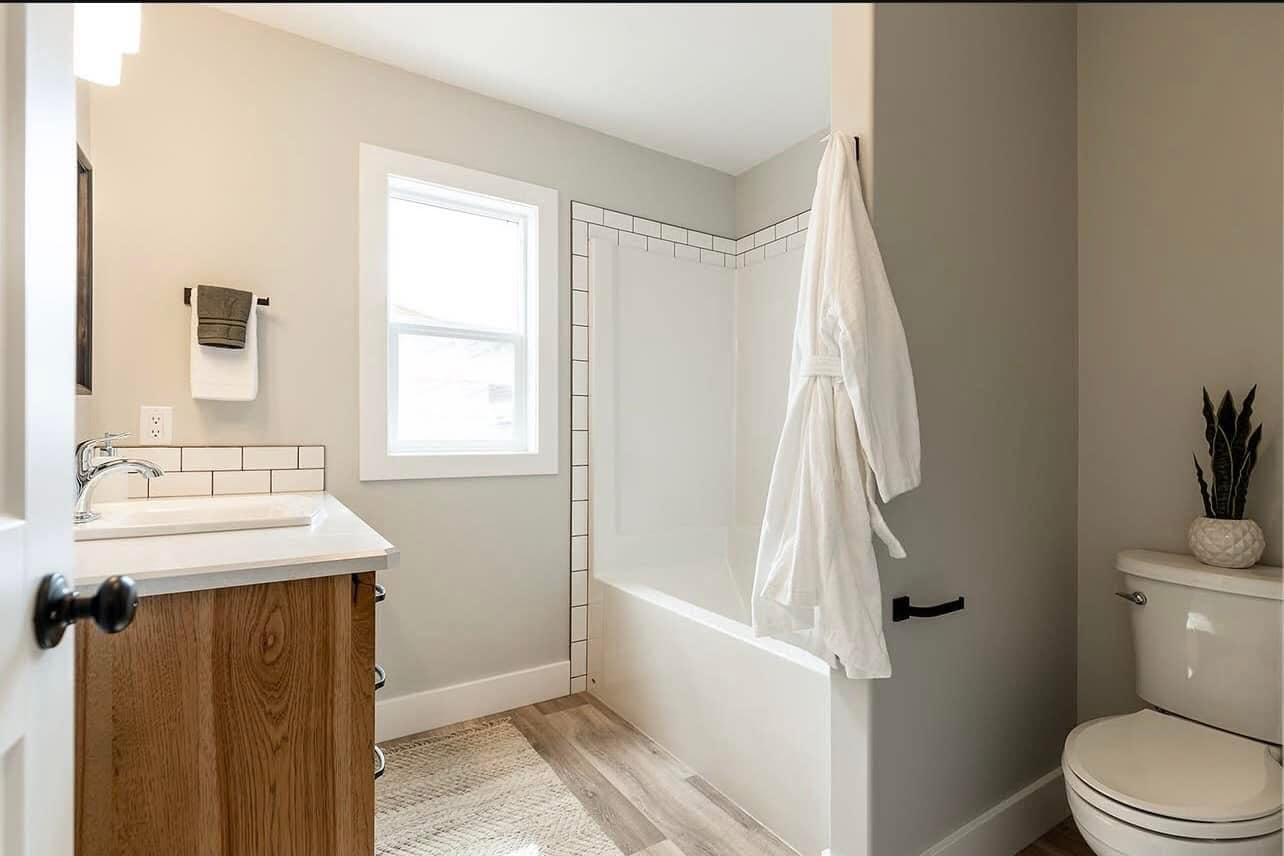 A modern bathroom with light gray walls features a bathtub with a white tile surround, a wooden vanity with a white sink, and a white robe on a hook. A plant sits on the toilet, adding a touch of greenery.