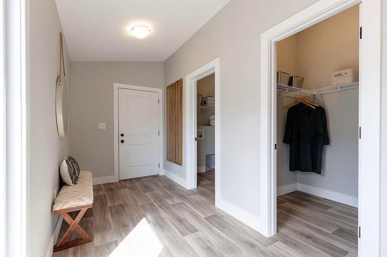 Bright entryway with light wood flooring, white walls, and two open closets. A simple bench and round mirror add a touch of elegance and functionality.