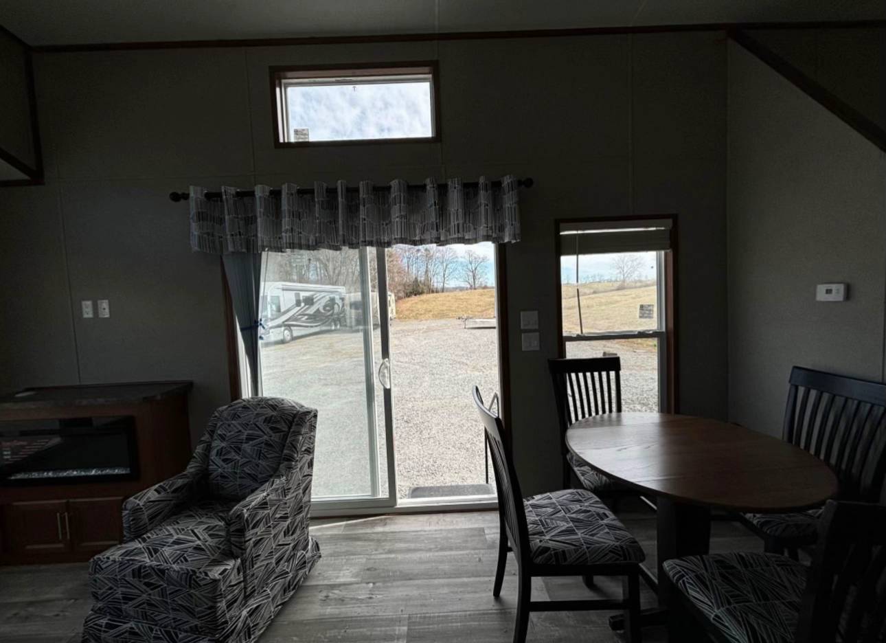 A cozy room with dim lighting features a patterned armchair, round wooden table, and chairs. A sliding glass door offers a view of a gravel path and grassy area. The room conveys a peaceful and inviting atmosphere.