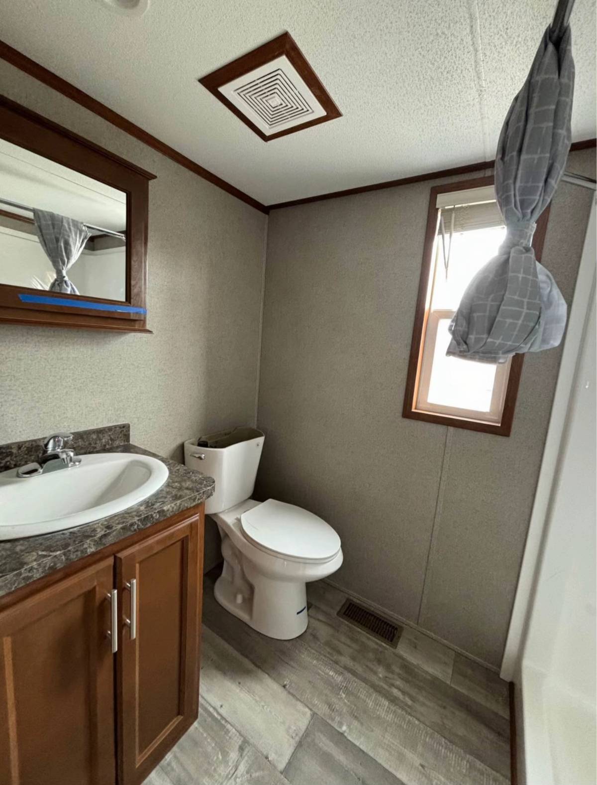 Compact bathroom features a sink with brown cabinets, a white toilet, and a window with a tied gray curtain. Modern decor and neutral tones are used.