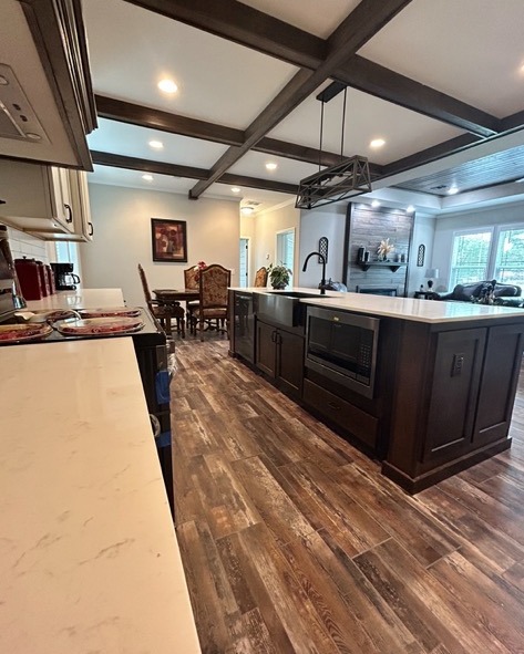 Spacious modern kitchen with dark wood island and light marble countertops. Features a dining area, wooden floor, coffered ceiling, and natural light.