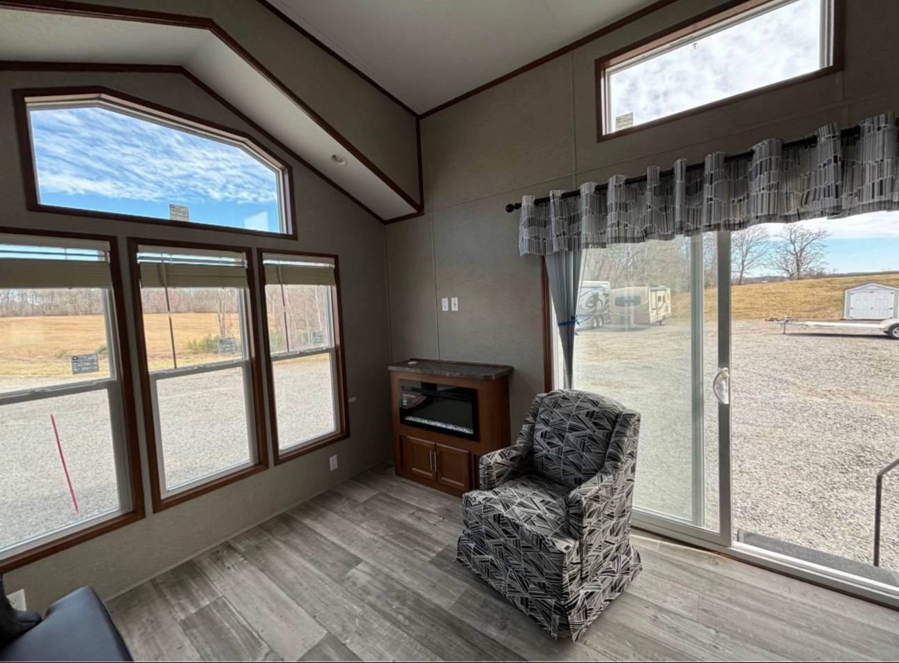 Cozy room with four large windows and sliding glass door showcasing a grassy field. Features an armchair, electric fireplace, and patterned curtains.