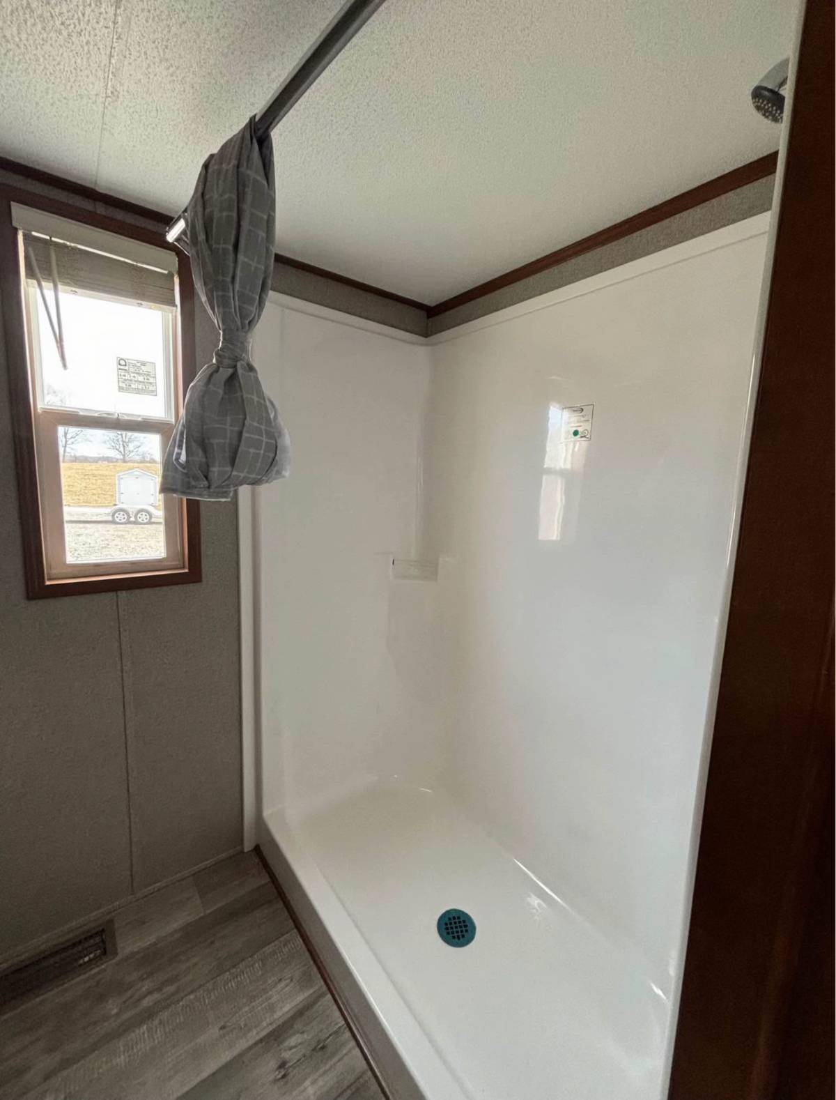 A simple white shower stall with a plaid shower curtain tied to the side is shown. Sunlight streams through a window, casting a soft glow.