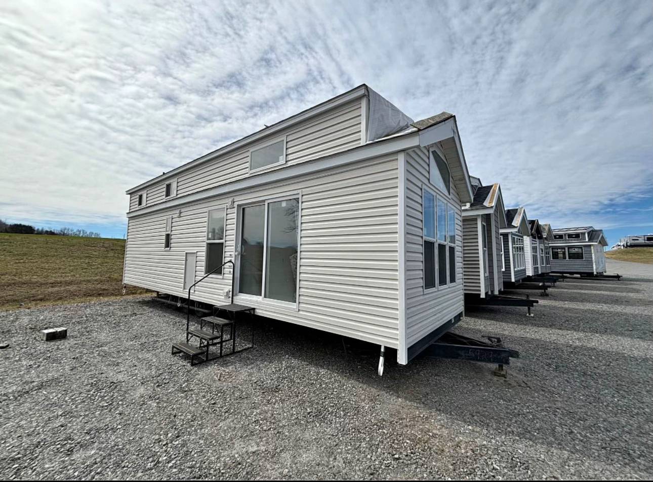 Row of modern white tiny houses on trailers in a gravel lot under a cloudy sky. Each house has large windows and simple metal steps. Quiet, open setting.