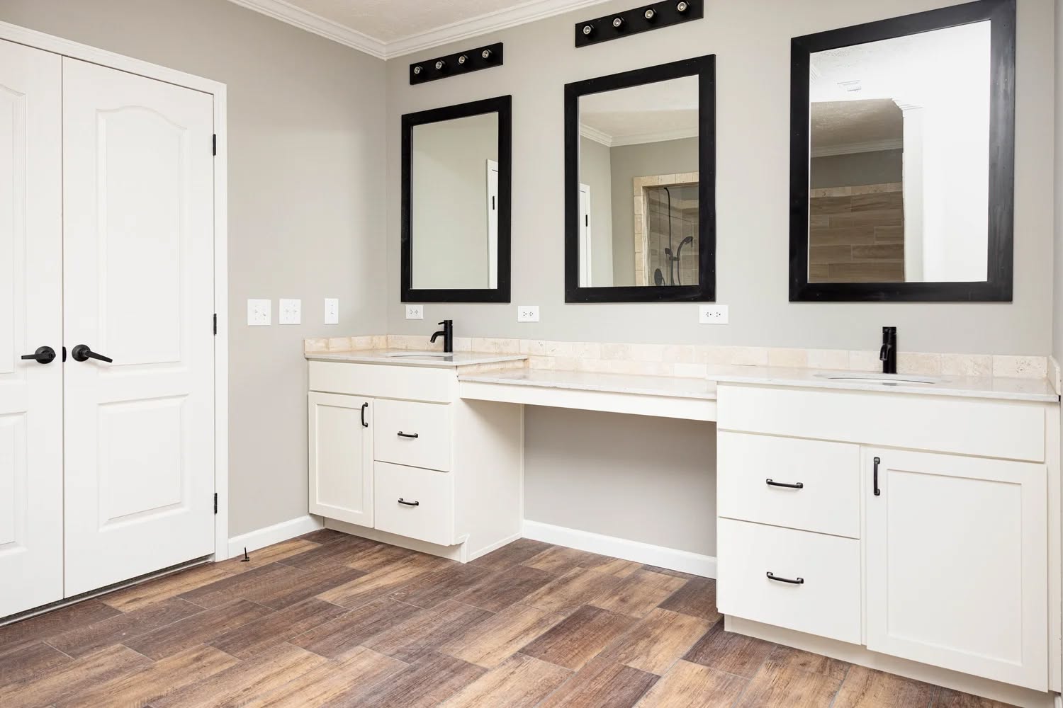 Bright bathroom with two white vanities, black faucets, and large mirrors. Wooden floor, neutral walls, and double doors create a modern, clean look.