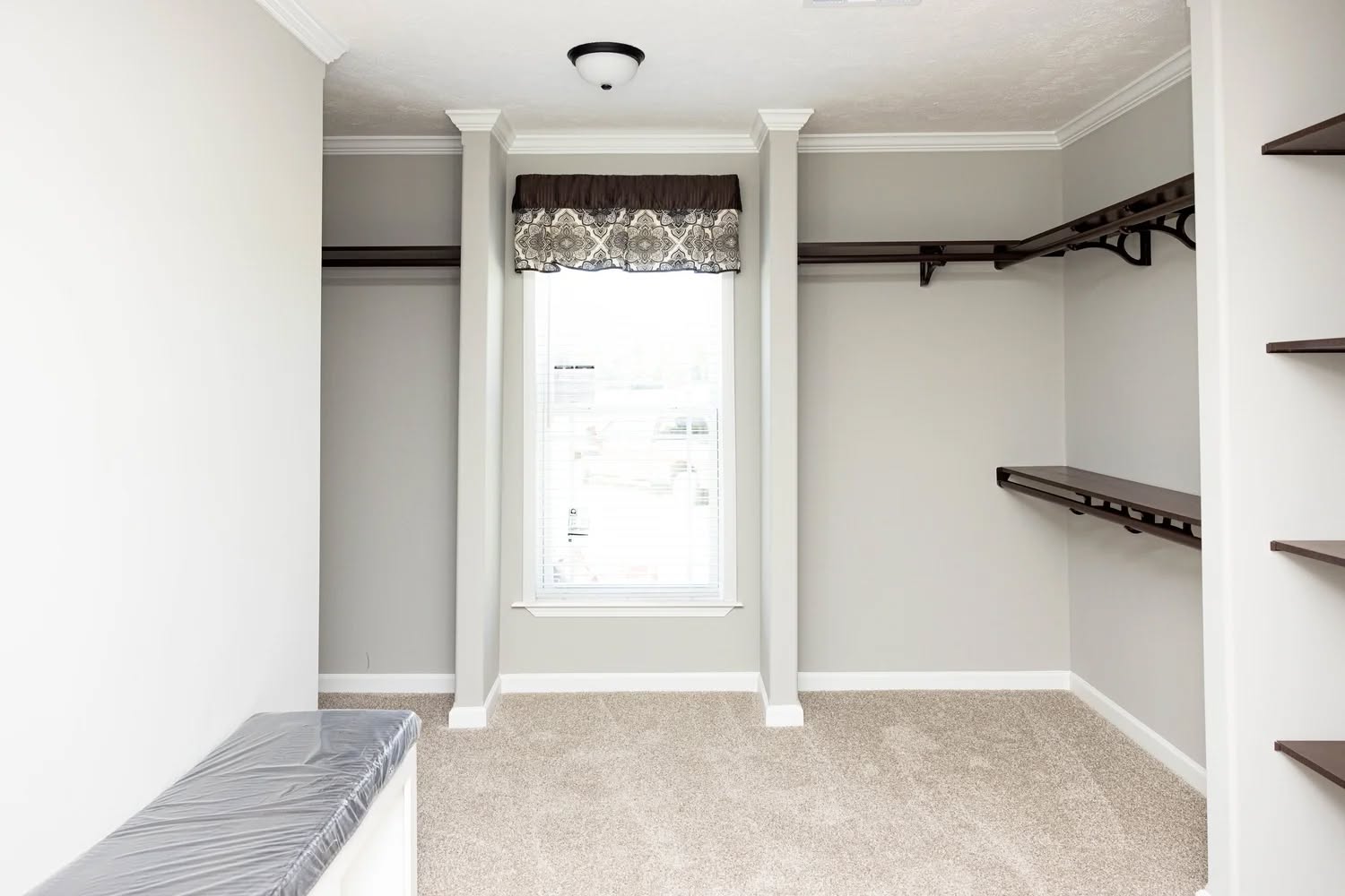 Spacious walk-in closet with beige carpet, light gray walls, and a large window with a patterned brown valance. Wooden shelves and rods are on the right.