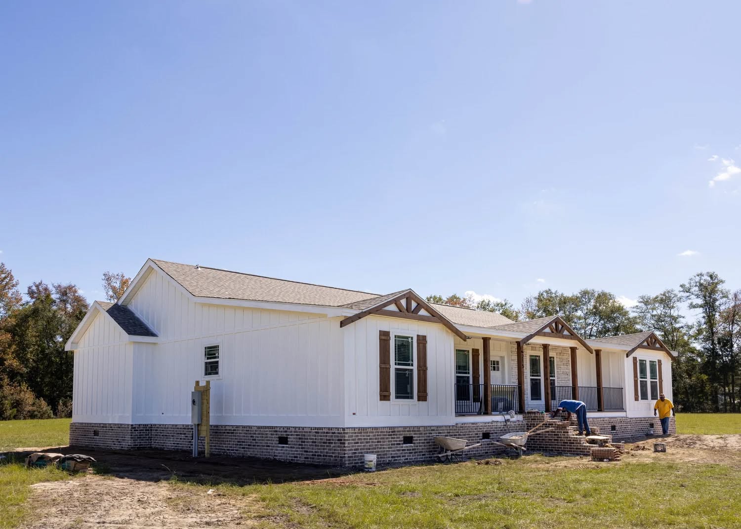 A single-story white house with a brick foundation sits in a grassy area under a clear blue sky. Two people work on the front steps, creating a sense of activity.