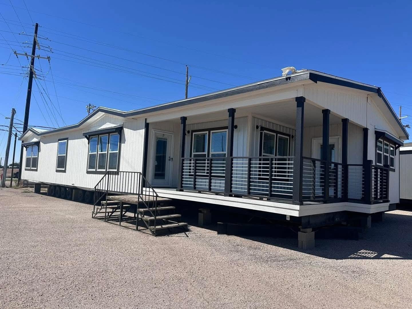 A white manufactured home with black trim sits on gravel under a clear blue sky. It features a small porch with metal railings and stairs leading up.