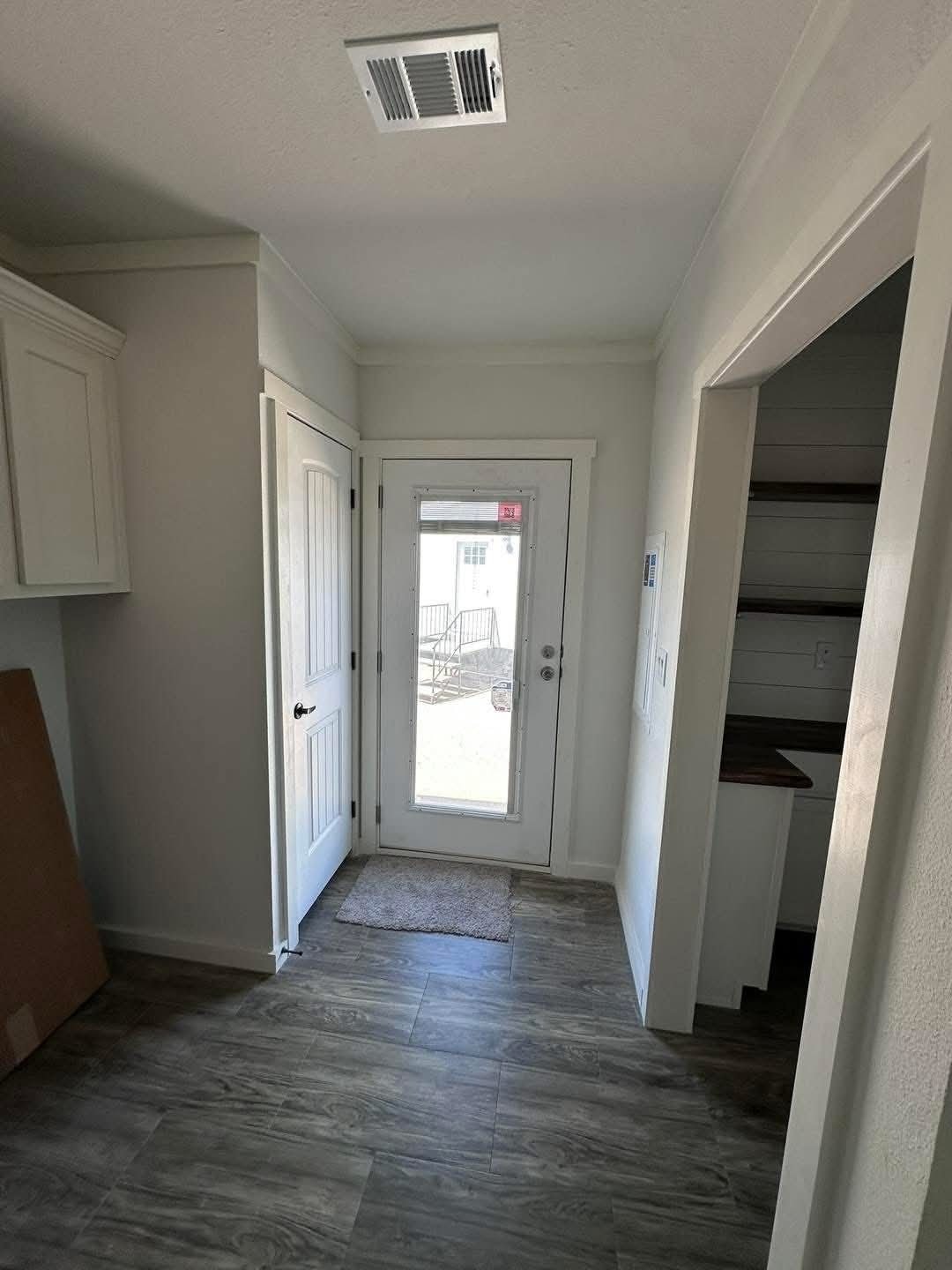 A bright entryway with a glass door leading outside, featuring wood-look floor tiles, white walls, and an open closet on the right. Airy and inviting.