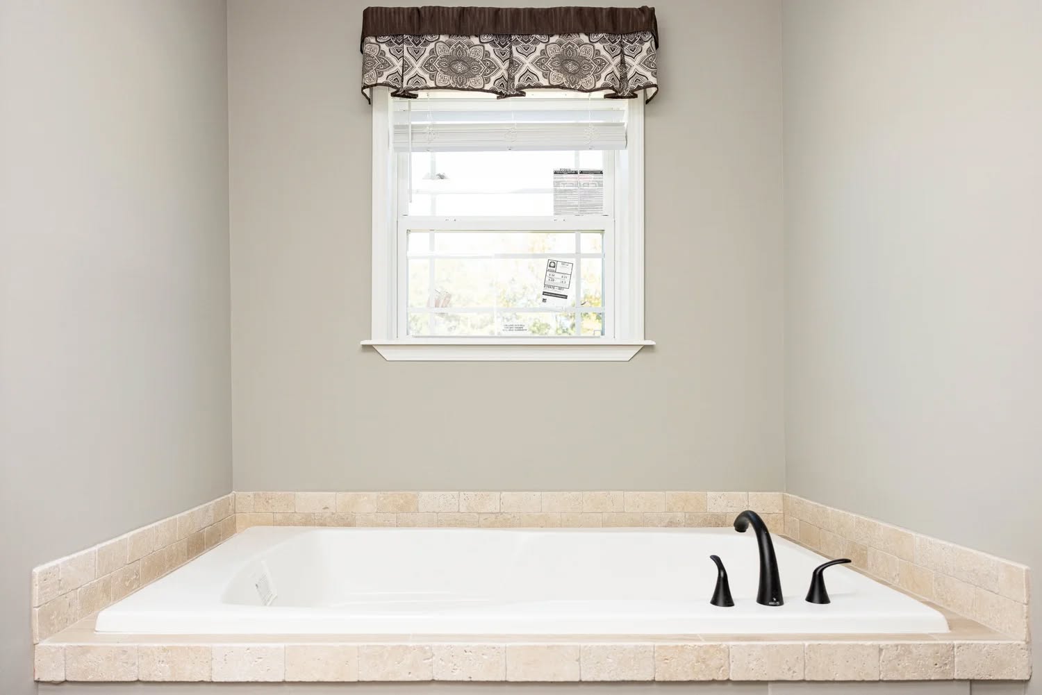 Spacious white bathtub set in beige tile with black faucets under a window featuring a patterned valance. The setting is serene and minimalistic.
