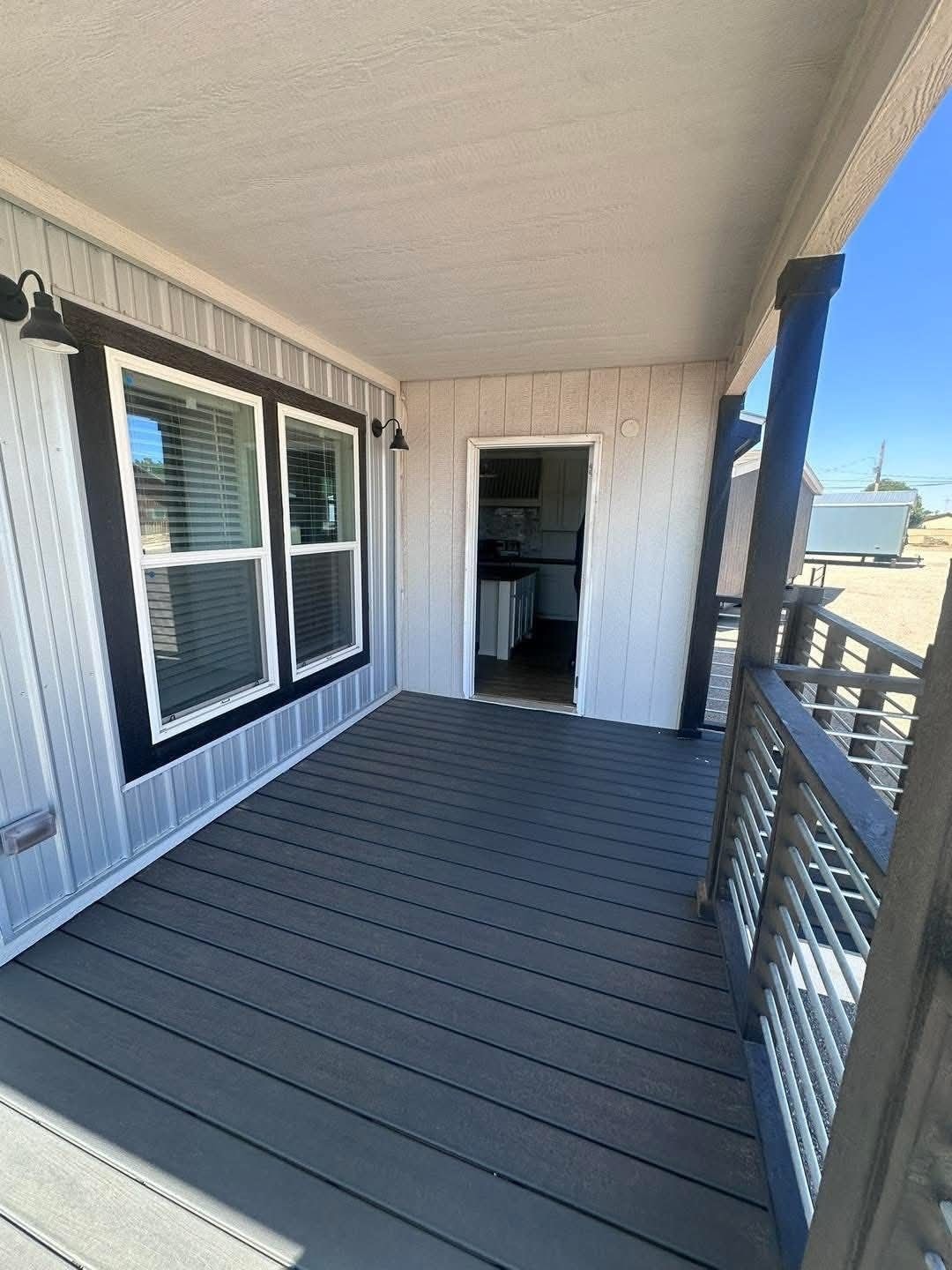 Covered porch with brown wooden decking, white siding, and two large windows on the left. An open door leads to an indoor kitchen. Bright, sunny day.