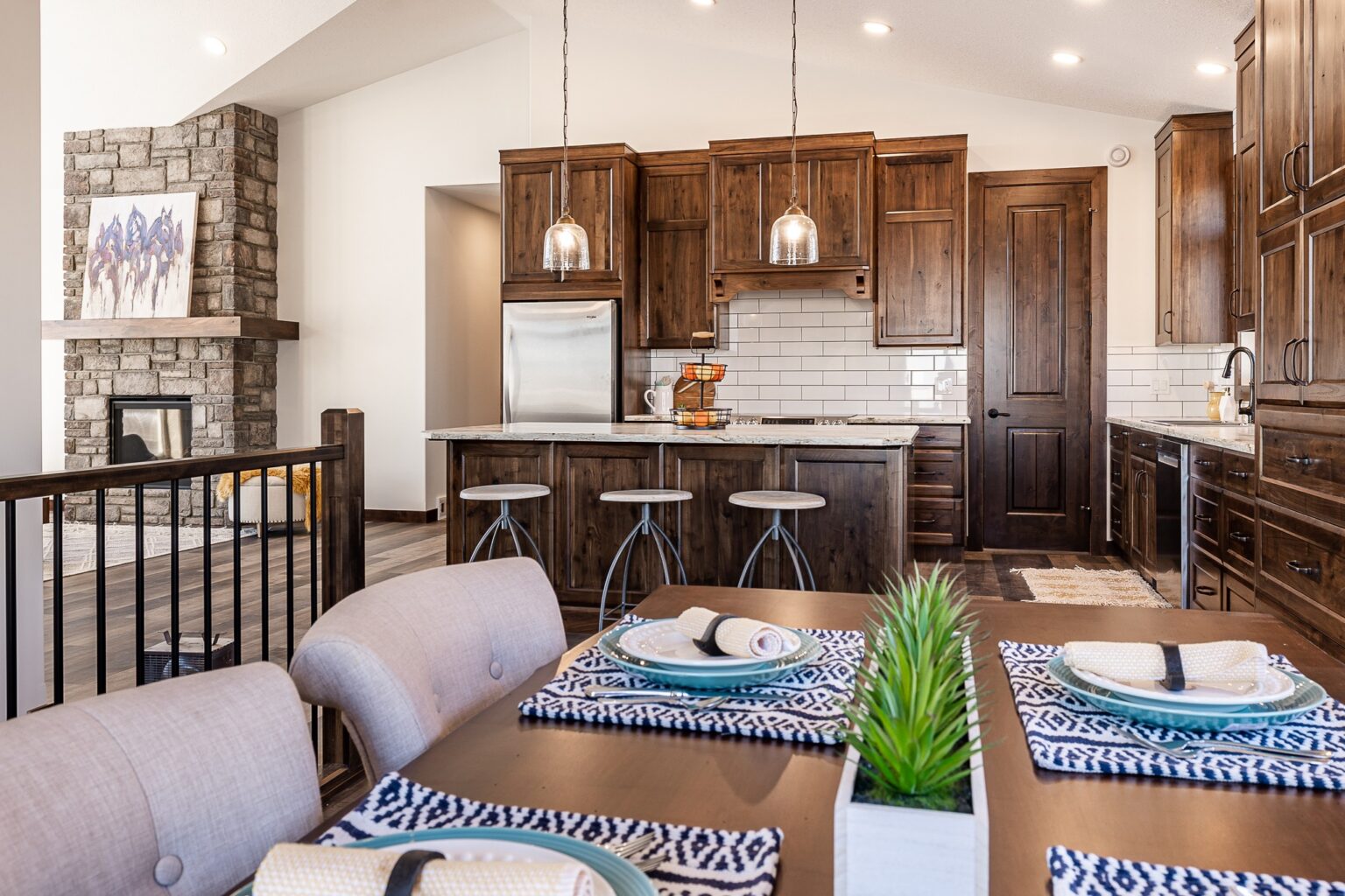Warm, rustic kitchen and dining area with wooden cabinets and island, metal stools, pendant lights, and a stone fireplace. Cozy and inviting ambiance.