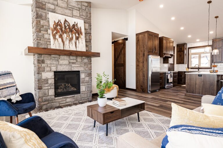 Modern living room with stone fireplace, wooden mantel, abstract art, and cozy navy chairs. Open kitchen with wood cabinets and pendant lights.