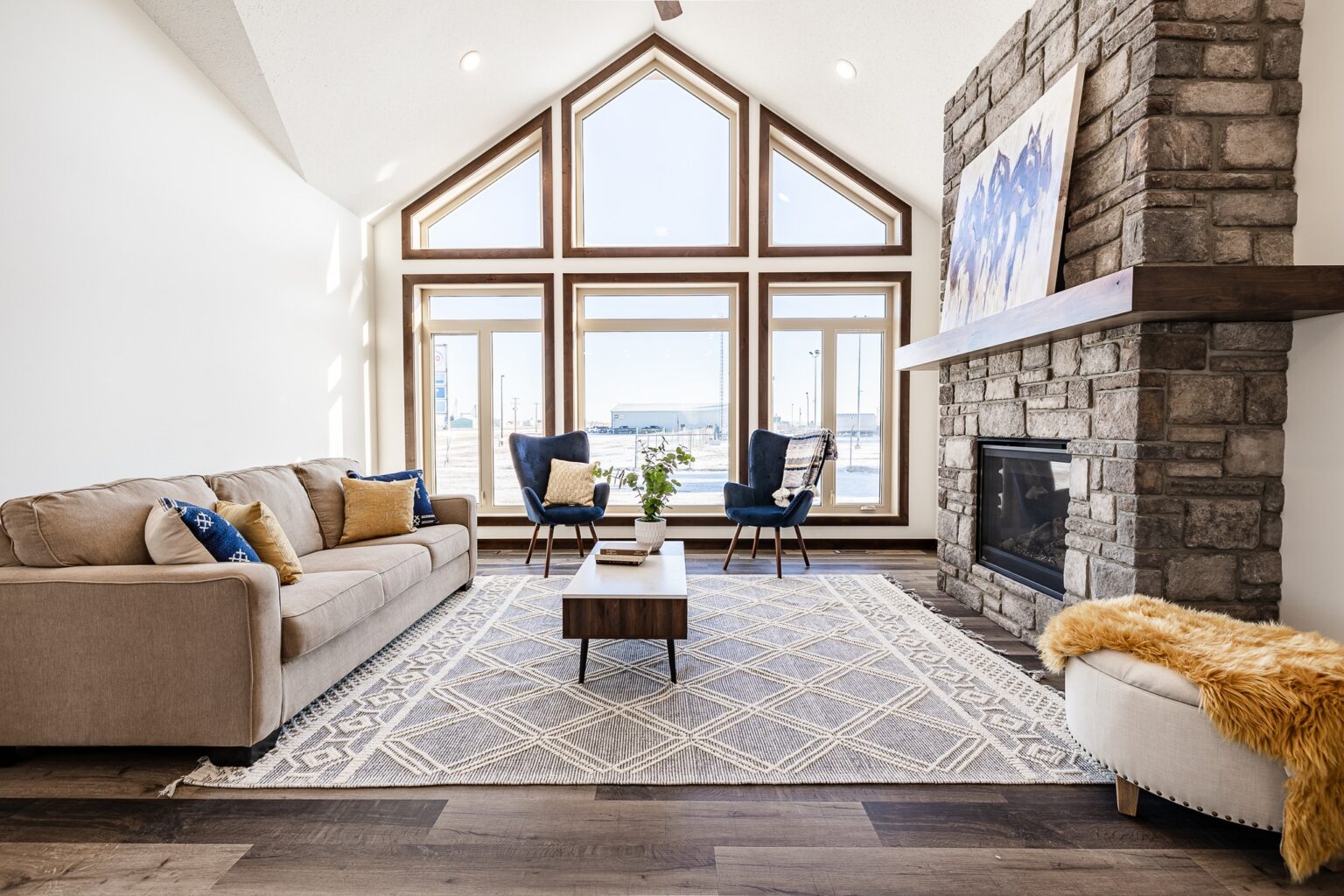Spacious living room with large triangular windows, beige sofa, blue chairs, stone fireplace, and geometric rug, conveying warmth and elegance.