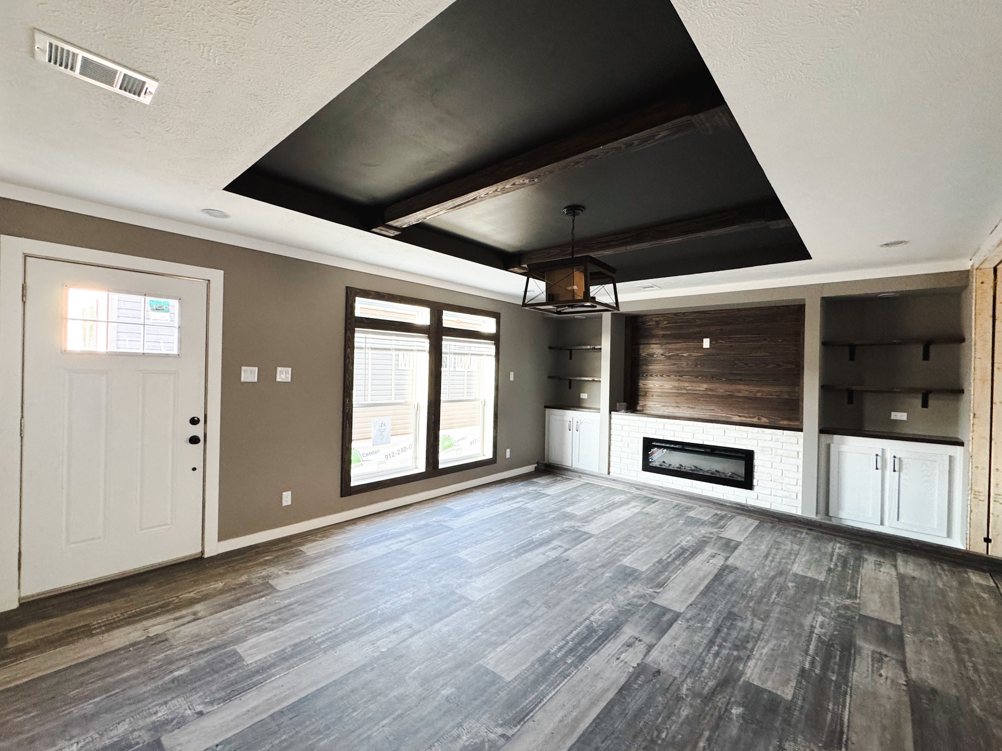 Modern living room with dark hardwood floors, a white door, and large windows. Features a black coffered ceiling, wood-paneled wall, and built-in fireplace.