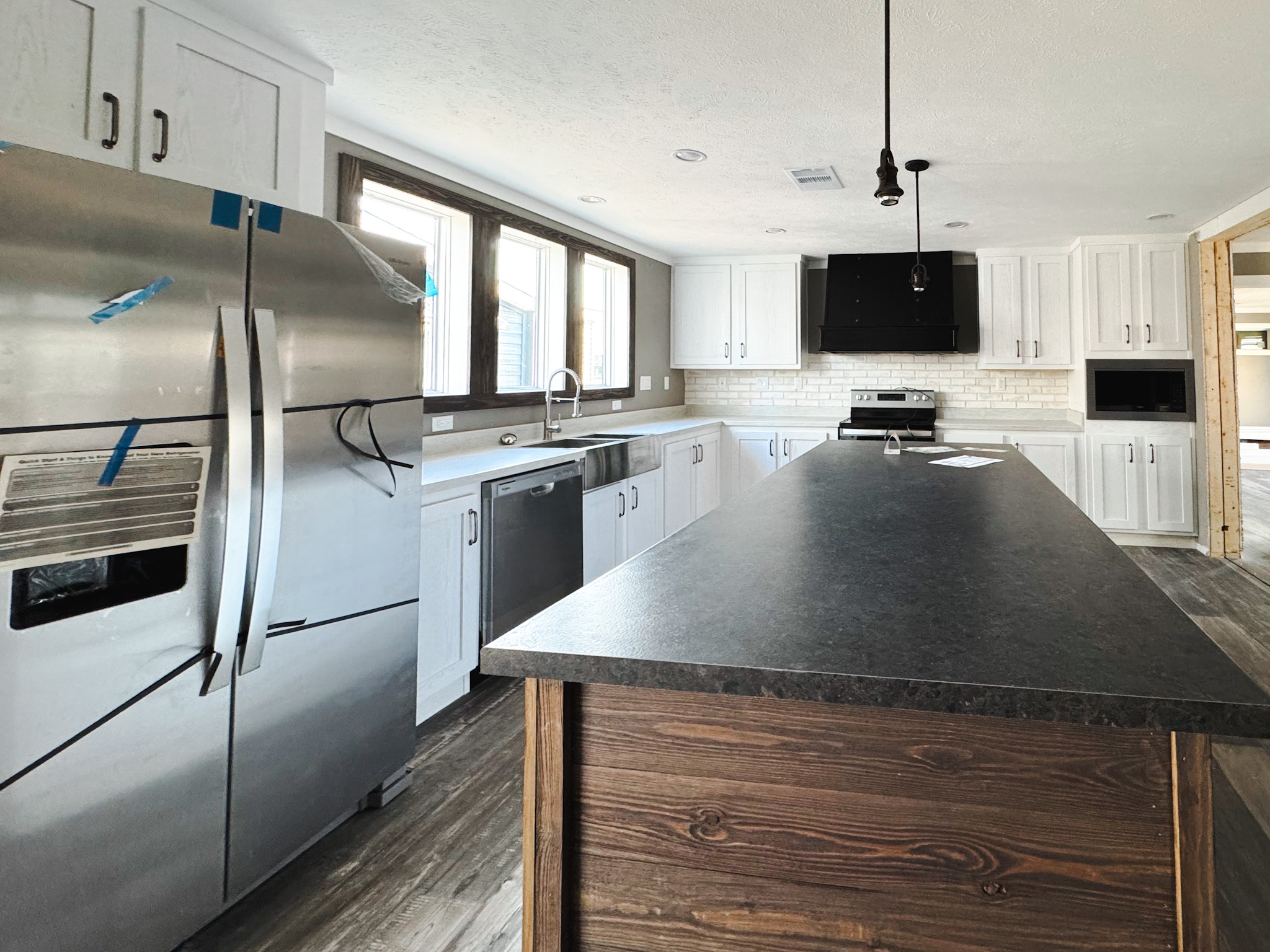 Modern kitchen with stainless steel appliances, a large center island, wood flooring, white cabinets, and a sleek black range hood under bright natural light.