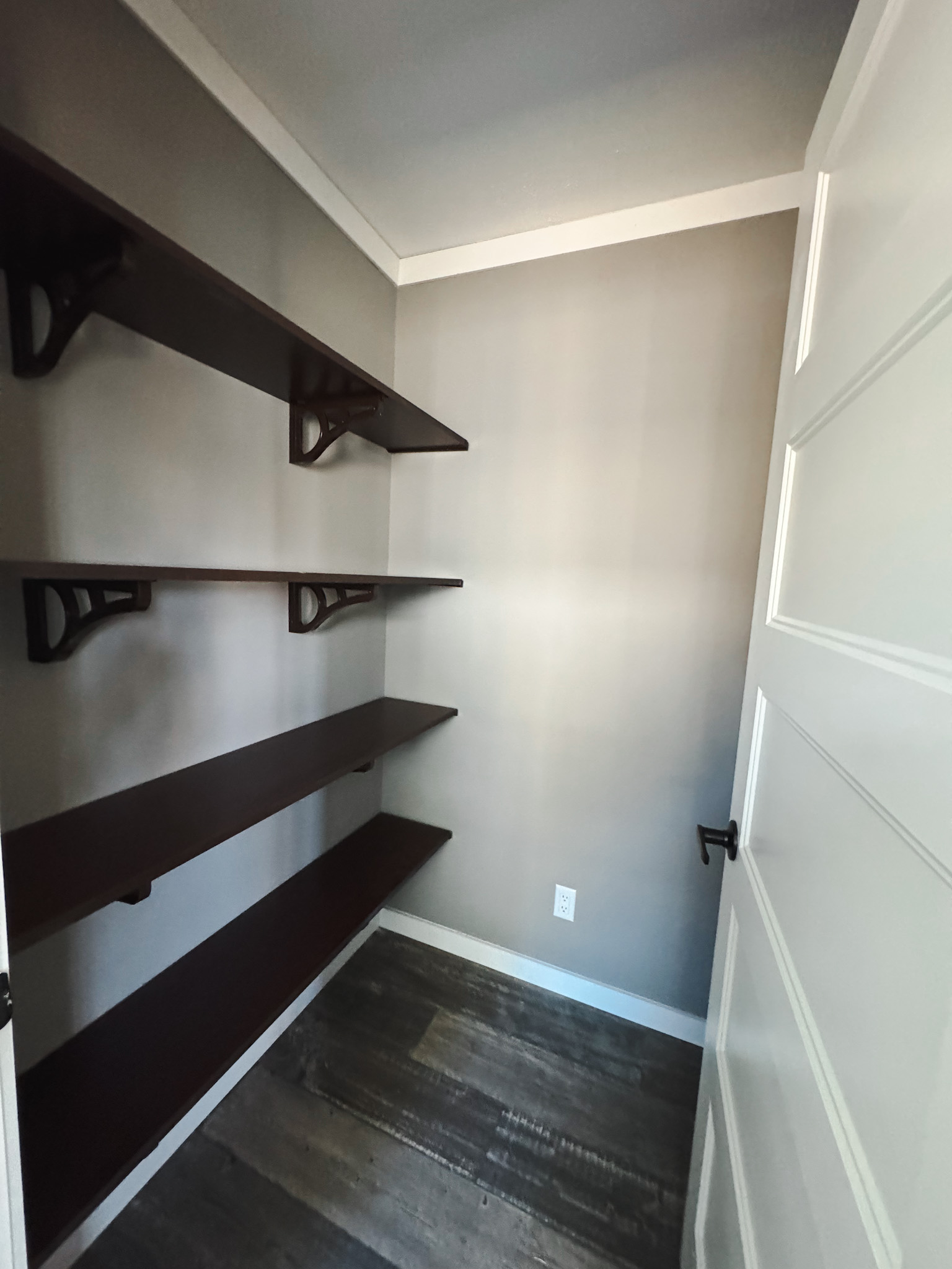 Small, empty closet with three dark wooden shelves on the left wall. Gray walls, white trim, and a brown wooden floor create a minimalist, clean atmosphere.