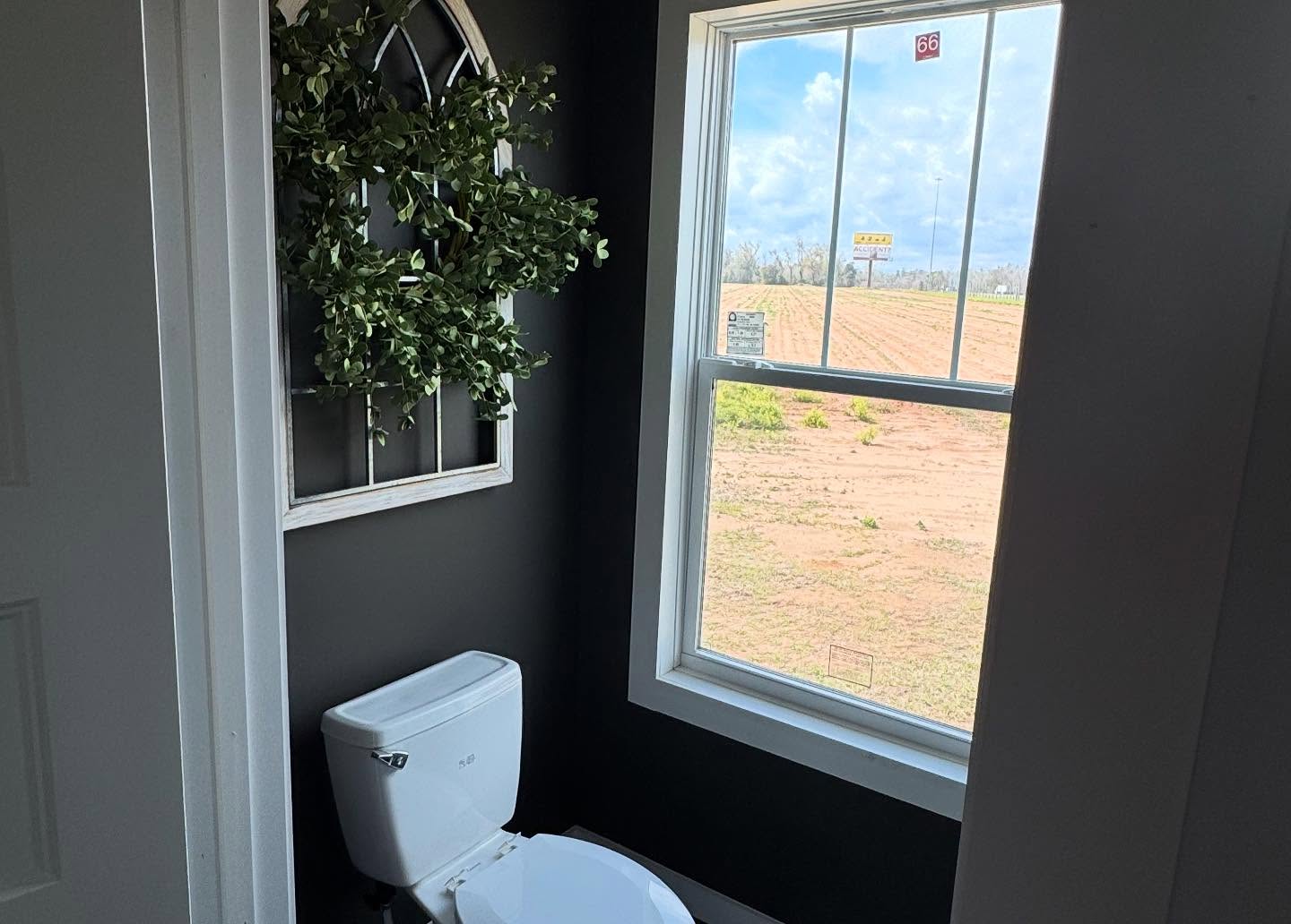 A cozy bathroom with a white toilet against a dark wall. A lush green wreath hangs on a decorative window-style frame. A large window reveals a sunny outdoor view.