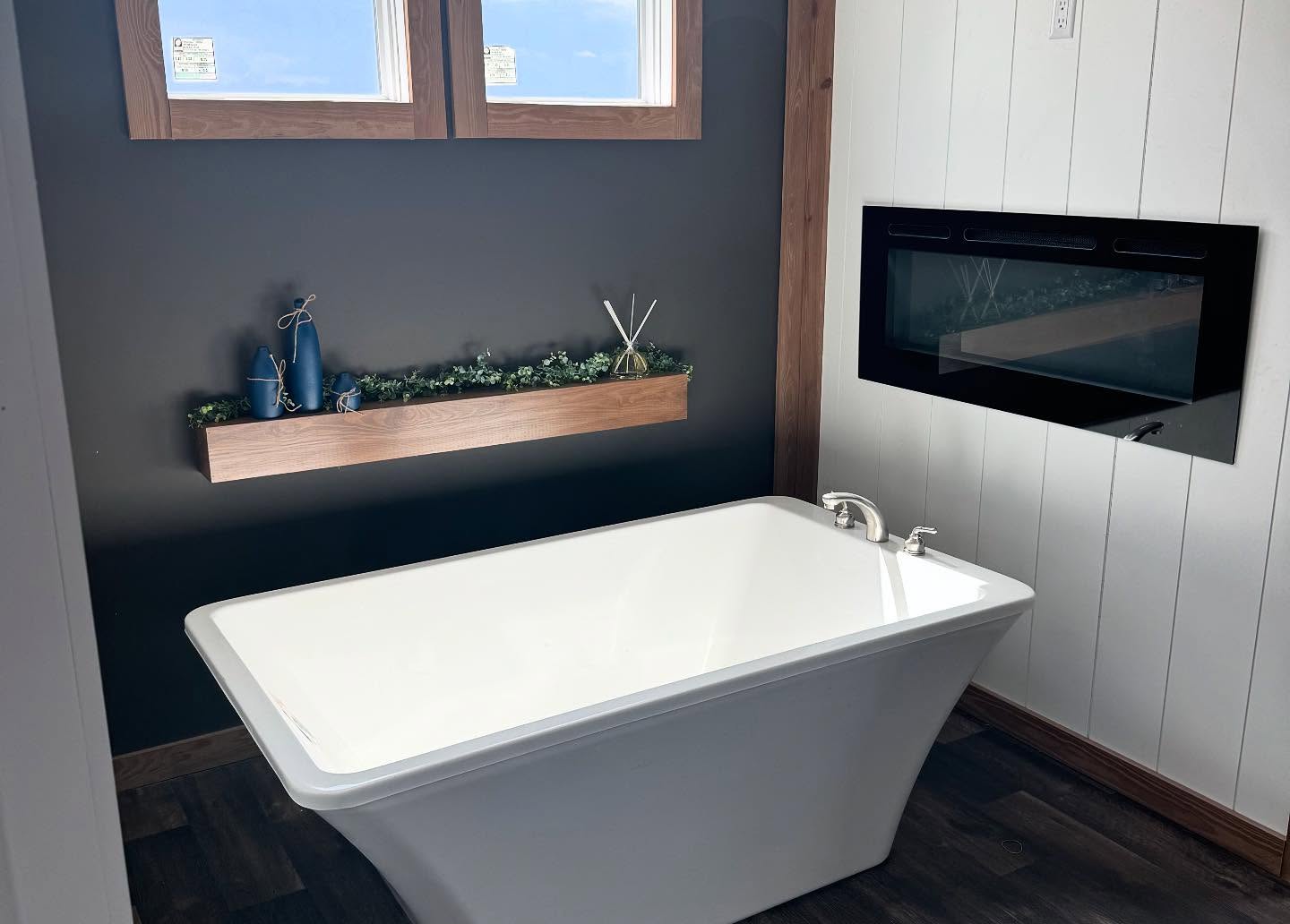 A modern bathroom with a white freestanding tub on dark wood flooring. A black accent wall features a floating shelf with greenery and blue decor. The room has a cozy and elegant atmosphere.