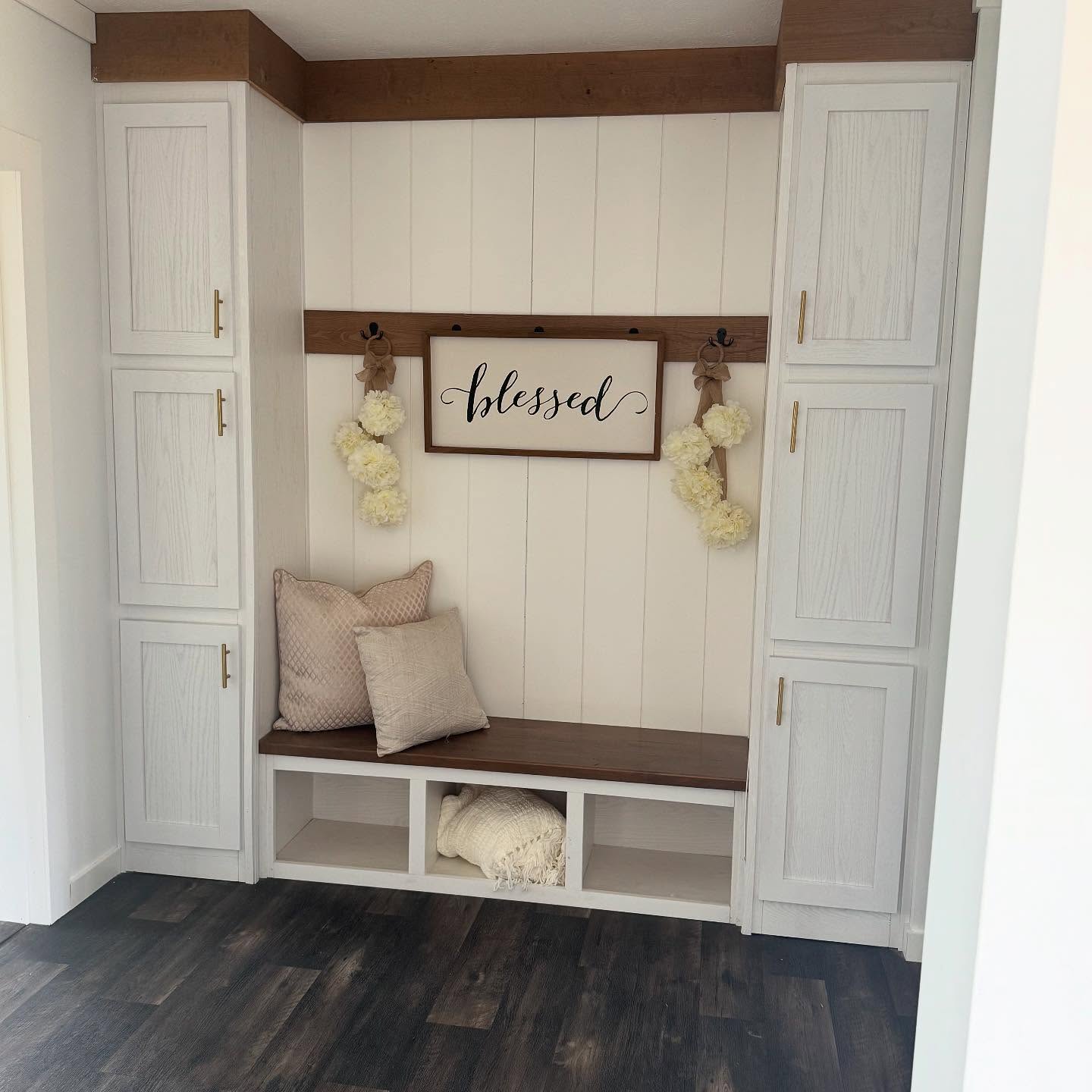 White entryway nook with a wooden bench, pillows, and blanket. "Blessed" sign and cream pom-pom garlands hang above. Cozy, welcoming atmosphere.