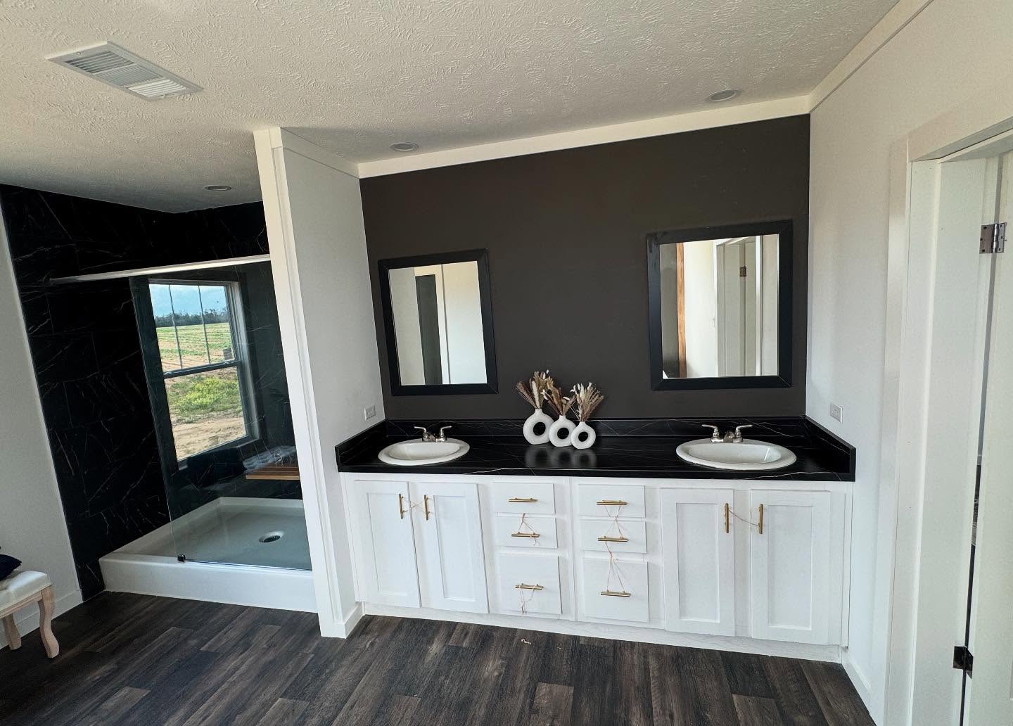 Modern bathroom with dual sinks, white cabinets, and black countertop. Two mirrors hang on a dark accent wall. A shower is visible on the left. Cozy and elegant.