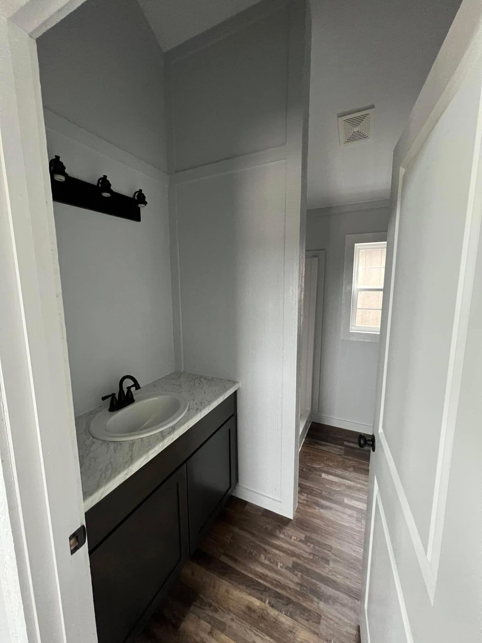 Narrow bathroom with white walls, dark wood floor, and a black vanity with a marble countertop. Single white window and modern light fixture above.