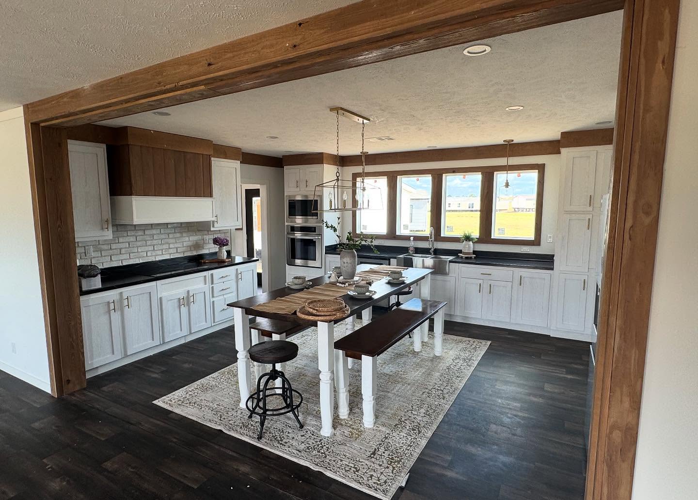 Spacious kitchen with white cabinets, dark countertops, and subway tile backsplash. Central wooden table and stools on a patterned rug. Warm, inviting tone.