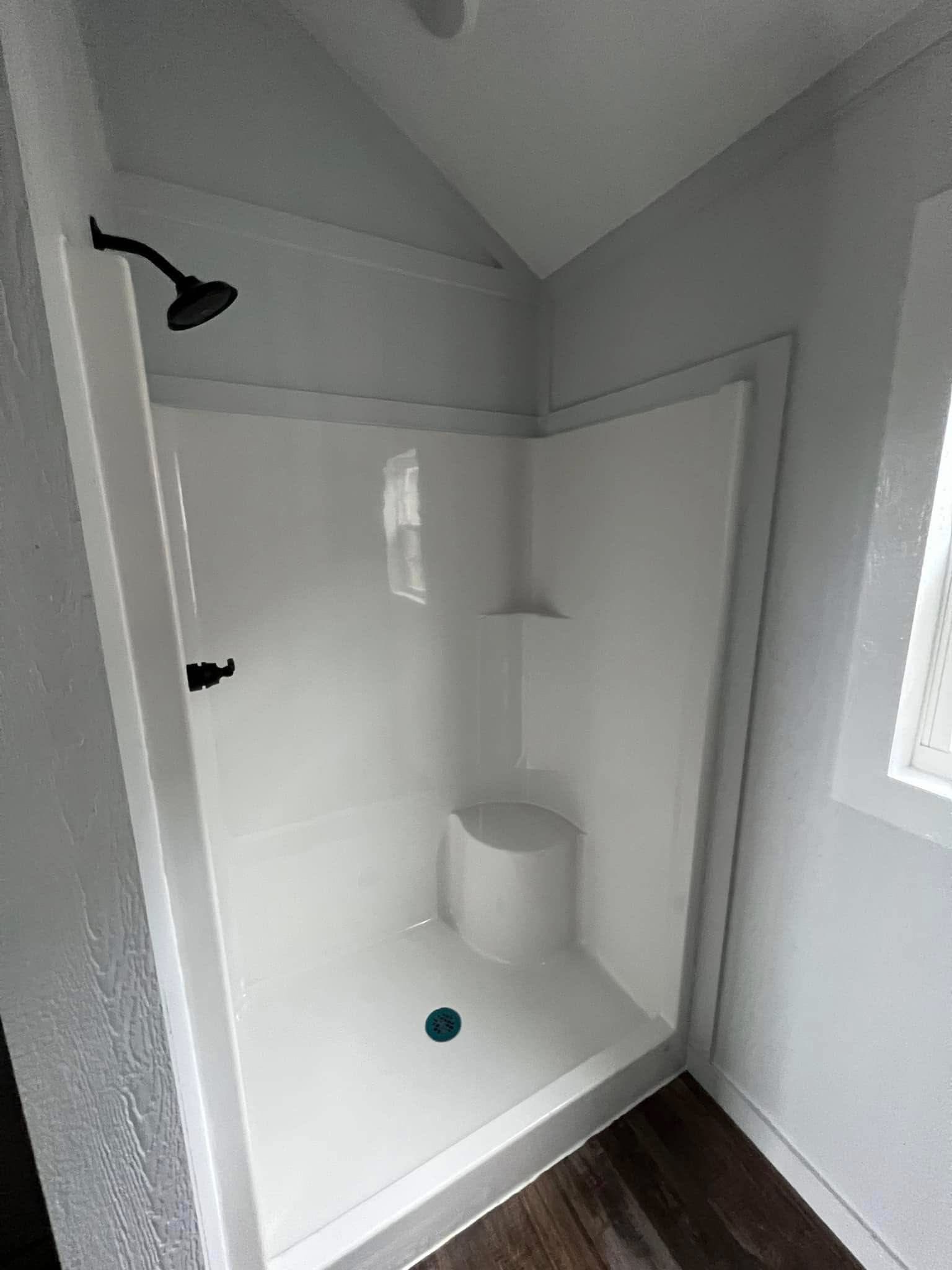 Simple shower stall with white walls, a black showerhead, and a teal drain. It has a built-in corner seat and shelf under a sloped ceiling, conveying a minimalist feel.