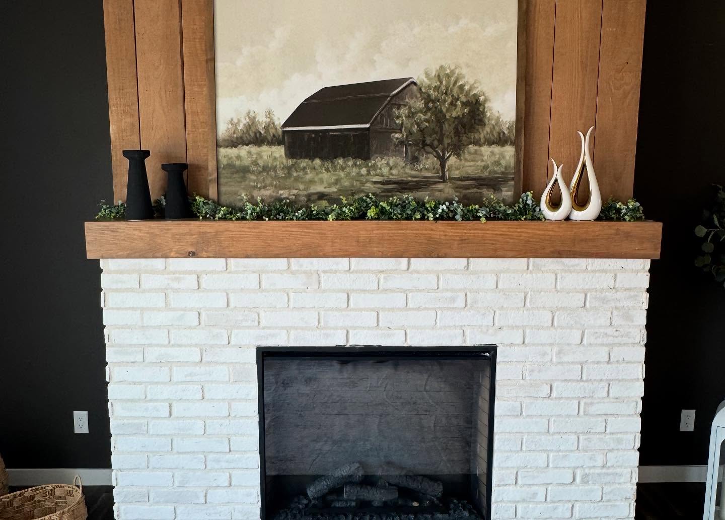 A white brick fireplace with a wooden mantel adorned with greenery, black vases, and white sculptures. A barn painting hangs above, creating a rustic, cozy vibe.