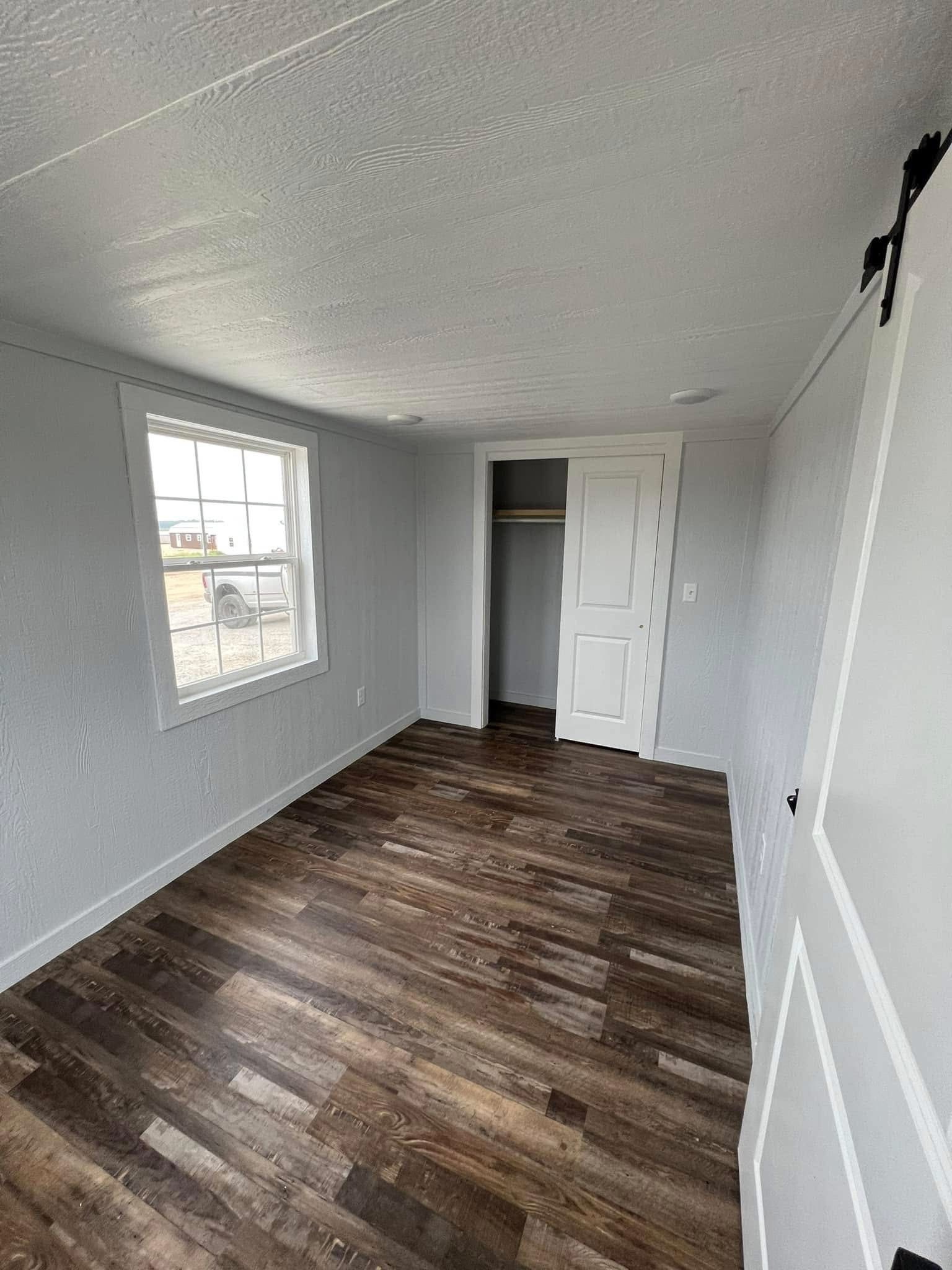 Small room with light gray walls and dark wood flooring. A large window on the left lets in natural light, and an empty closet is on the far wall.