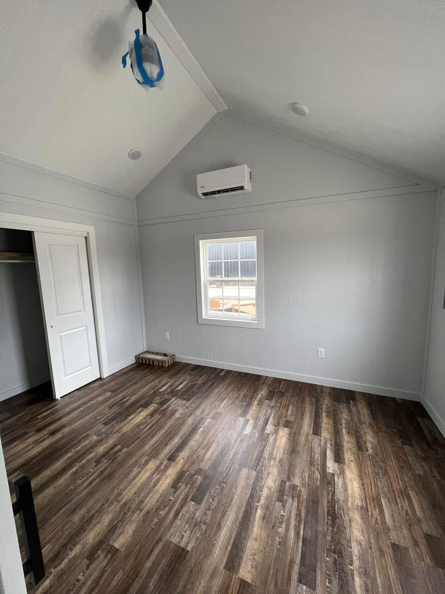 Empty room with light gray walls and vaulted ceiling. Wooden floor, small window, and a wall-mounted air conditioner. Door leads to a closet.