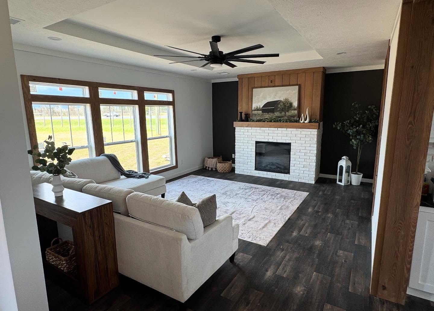 Modern living room with dark wood floors, white brick fireplace, and large windows with outdoor view. A beige sofa and soft rug create a cozy feel.