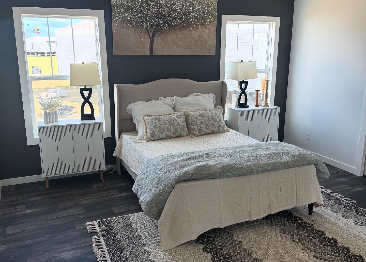 Modern bedroom with a gray accent wall featuring a tree painting above a beige bed. Two windows with light filtering through, flanked by white nightstands and lamps, add brightness. The space is cozy and inviting with a patterned rug.